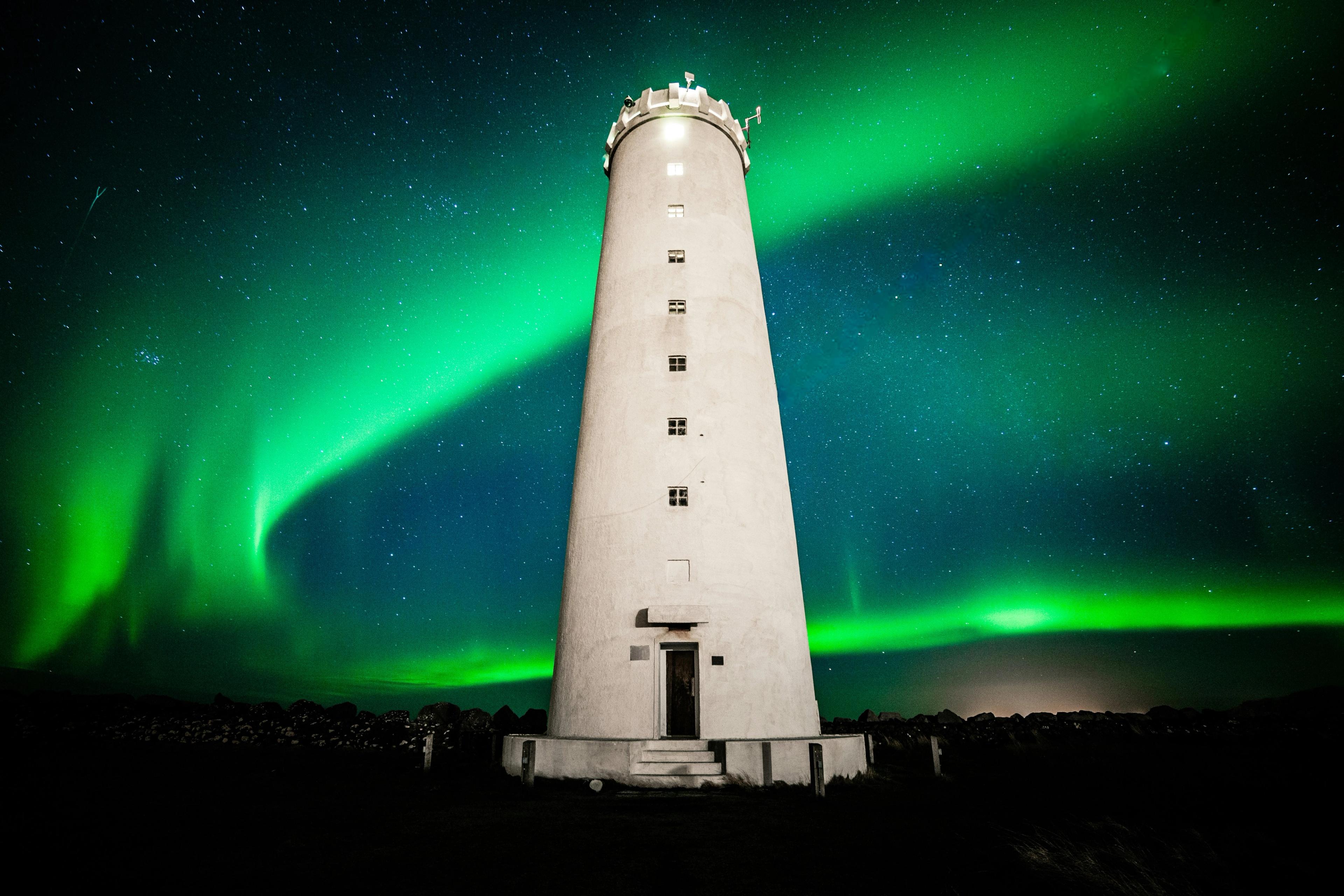 A tall white lighthouse at Grótta stands under a starry night sky illuminated by vibrant green Northern Lights.