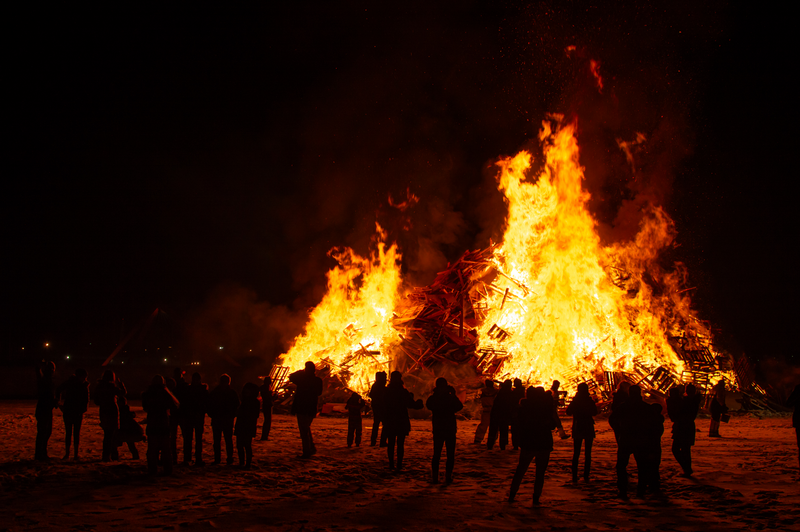 A group of people gathered around a large bonfire at night, watching the flames rise into the dark sky.