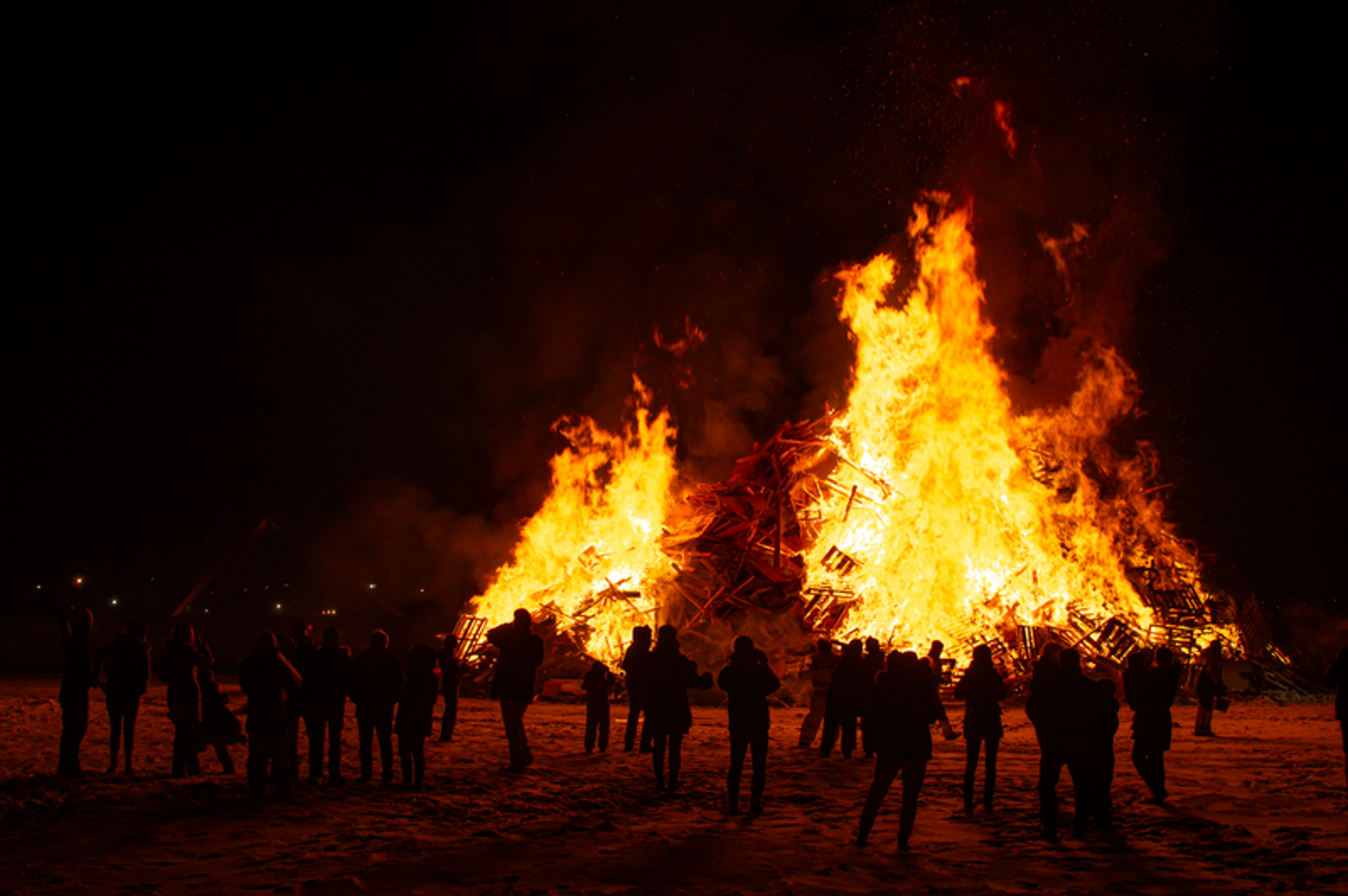 A group of people gathered around a large bonfire at night, watching the flames rise into the dark sky.