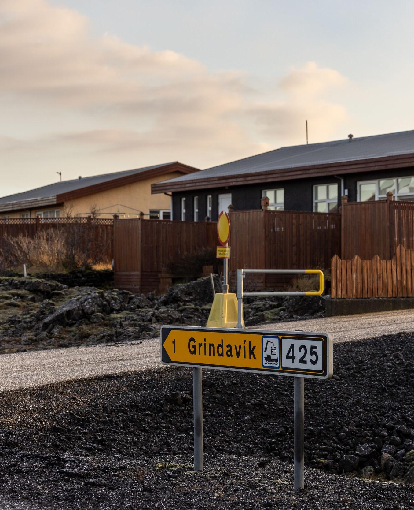 A road sign in a rural area points toward Grindavik on Route 425, with a backdrop of modest buildings and a cloudy sky. The scene captures the quiet, rugged landscape common to Icelandic towns.