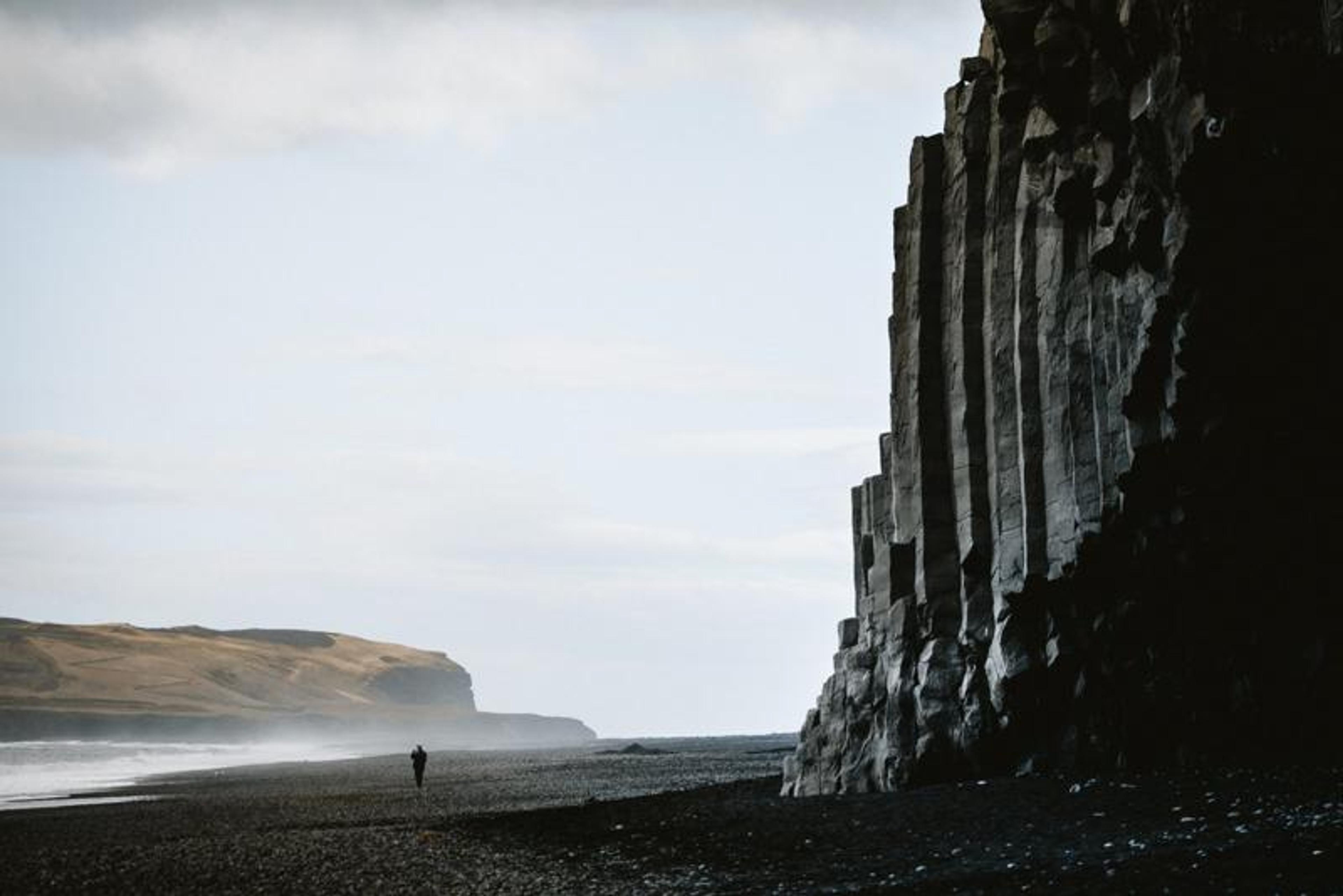 A lone figure walks on a black sand beach beside towering basalt columns, with misty waves and distant hills under a cloudy sky.