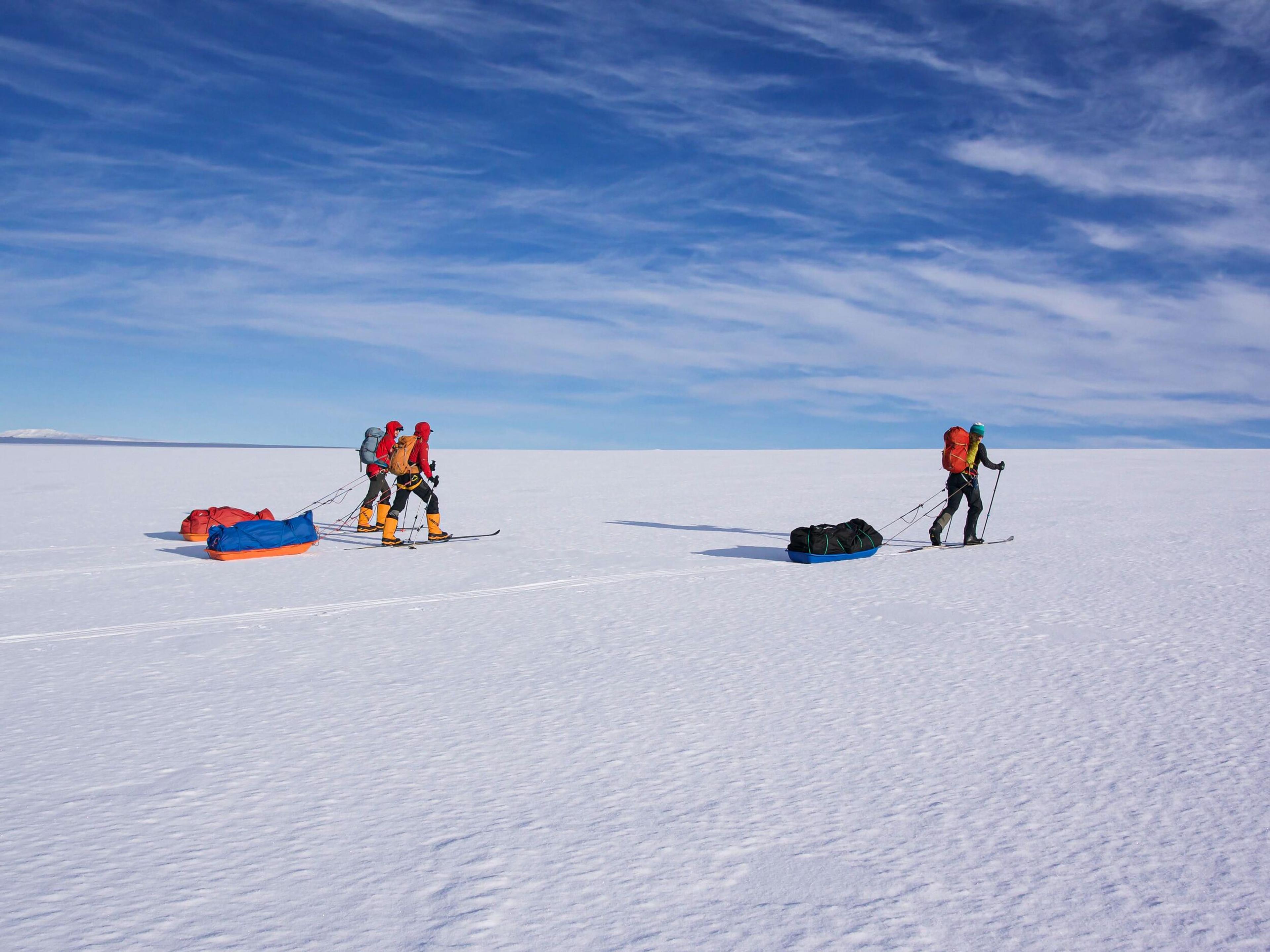 Explorers in bright gear cross an expansive snowy landscape, pulling sleds under a vast blue sky.