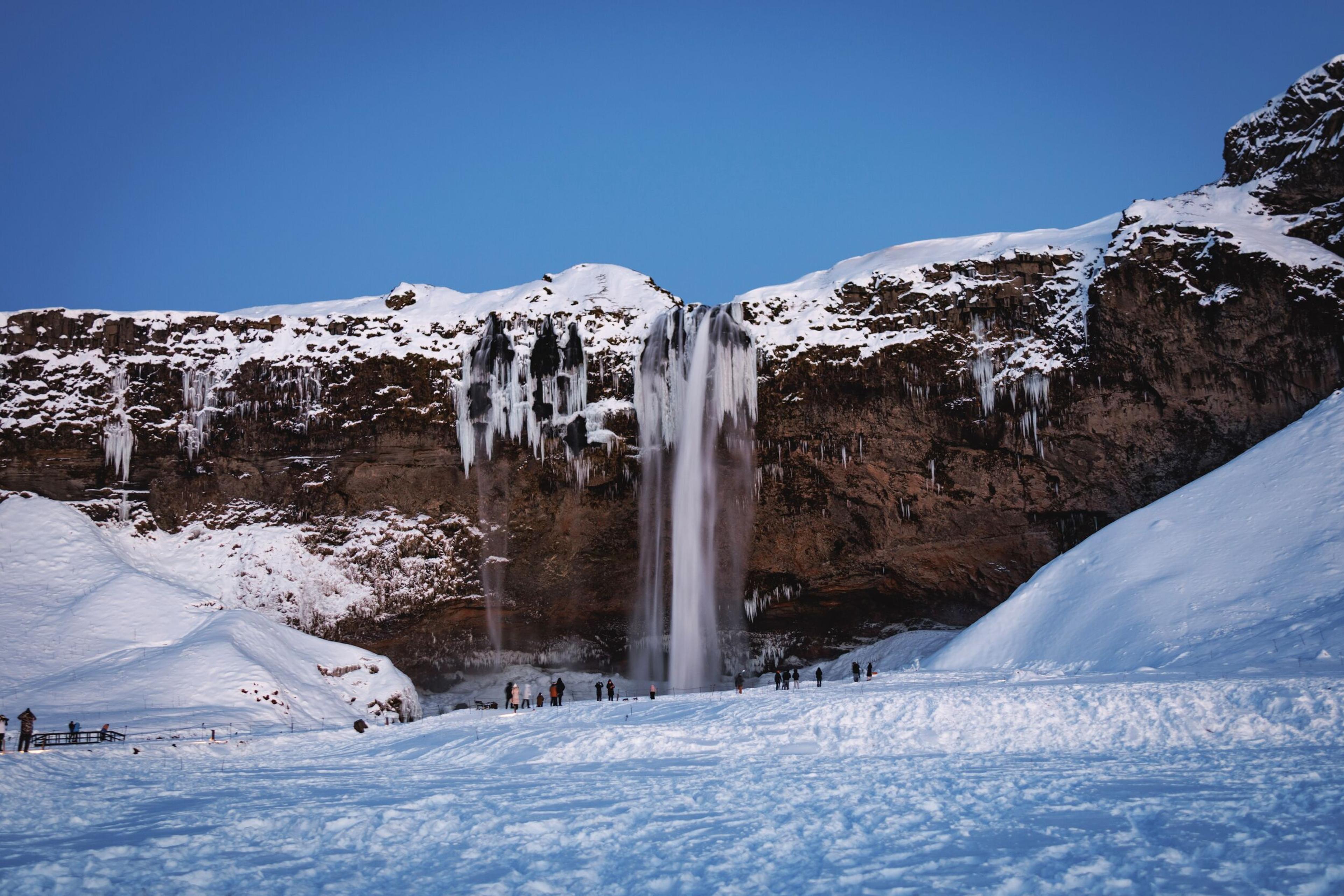 A waterfall plunges over a snow-covered cliff with prominent icicles, with visitors standing on the snowy ground below.