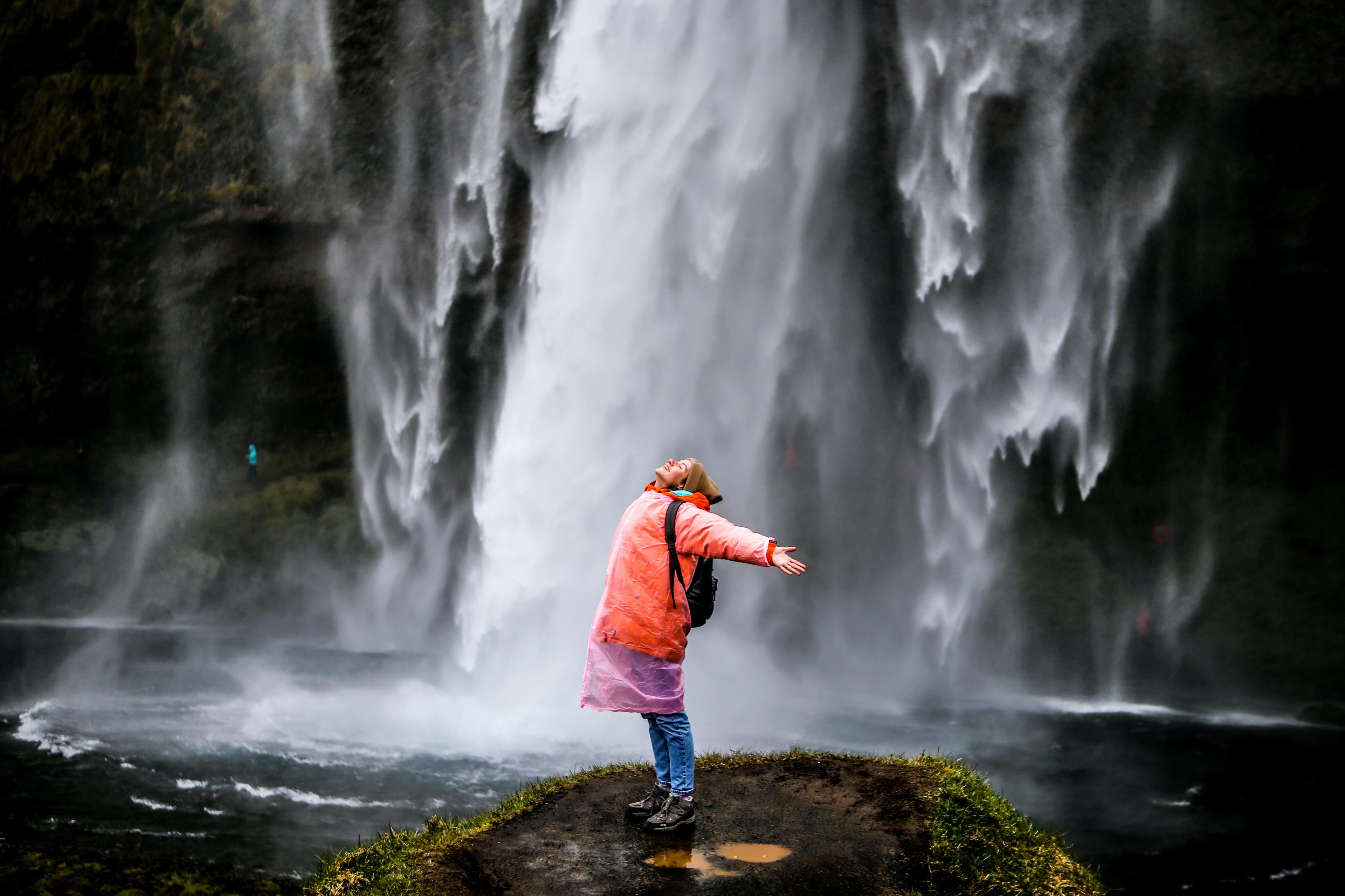 A person in a pink plastic raincoat joyfully spreading their arms beside the majestic Seljalandsfoss waterfall.