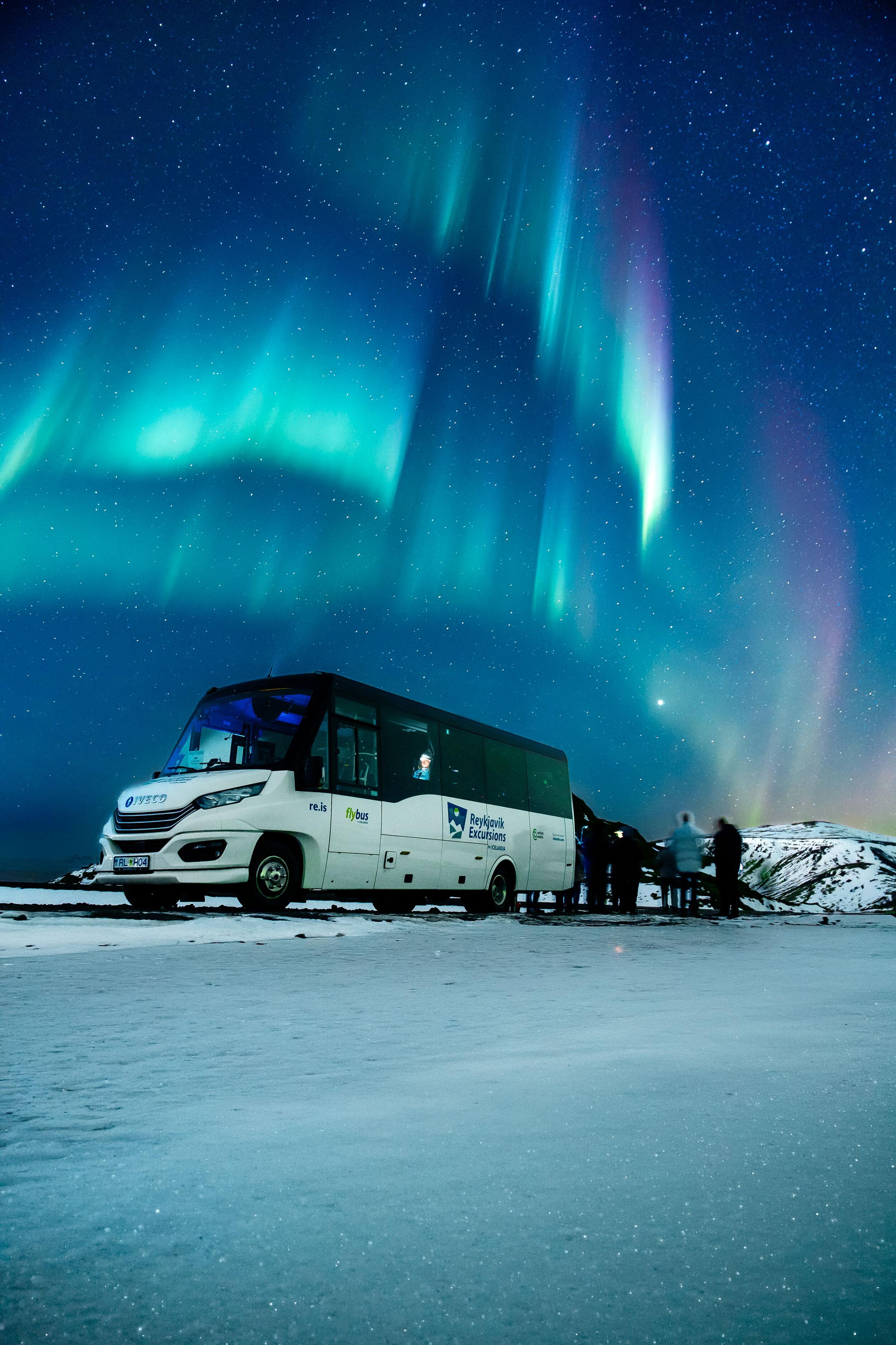 Reykjavík Excursions bus parked on a snowy landscape under vivid green and purple Northern Lights.