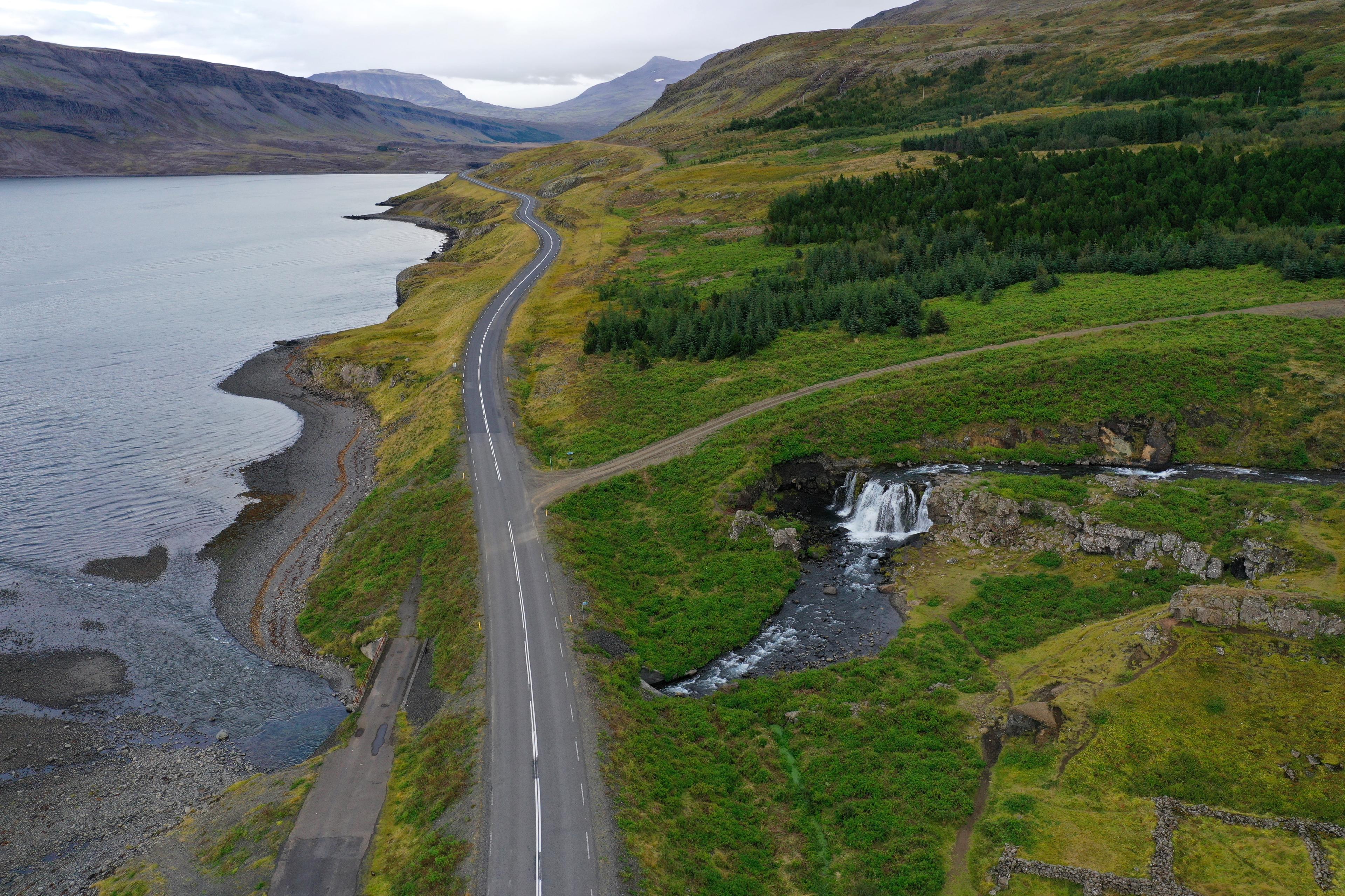 An aerial view reveals a serene Hvalfjörður, where a solitary road hugs the fjord's curve, bordered by lush green meadows and a quaint waterfall tumbling into the tranquil summer waters.