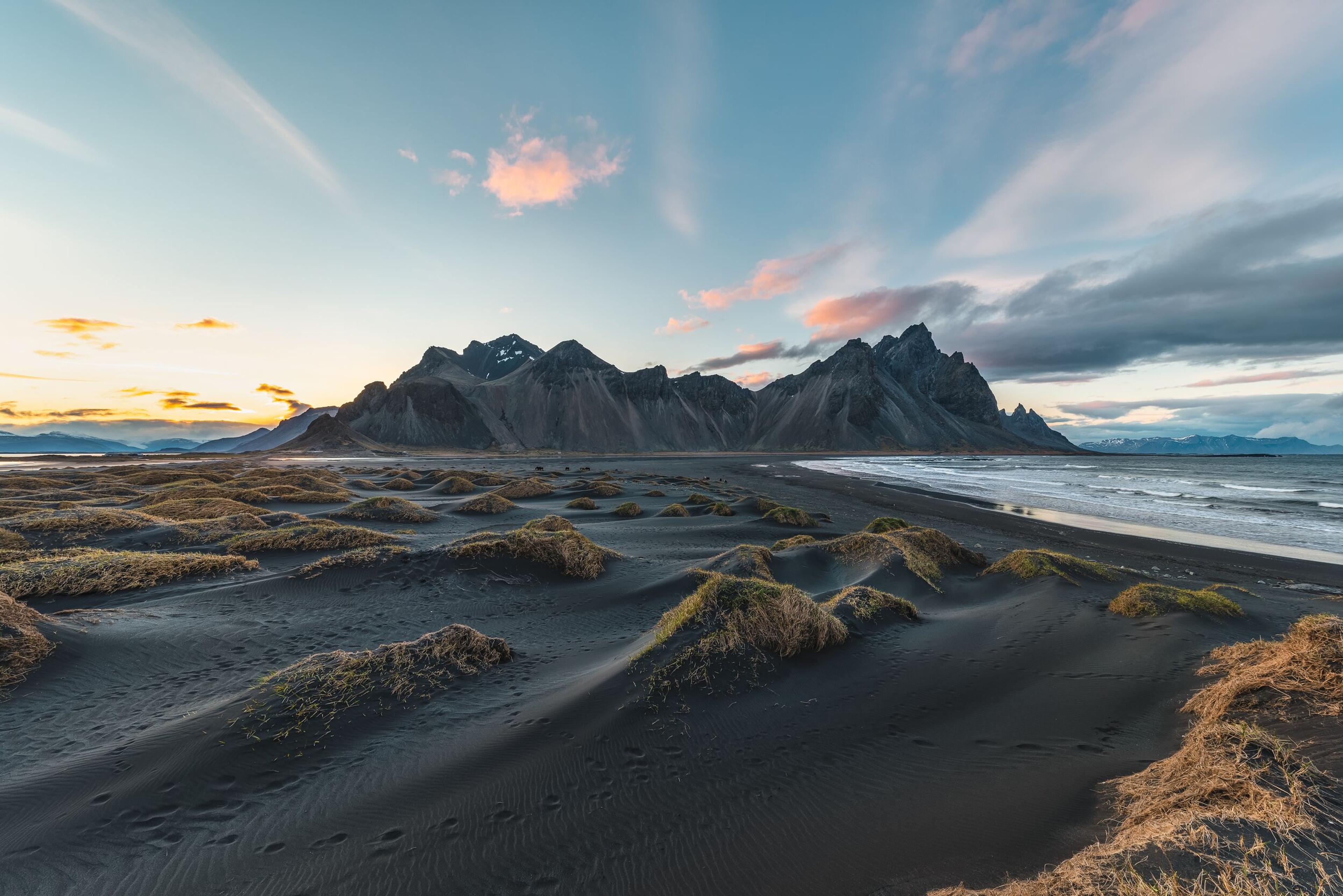 Black sand beach with grassy dunes, jagged mountains, and a colorful sunset sky.