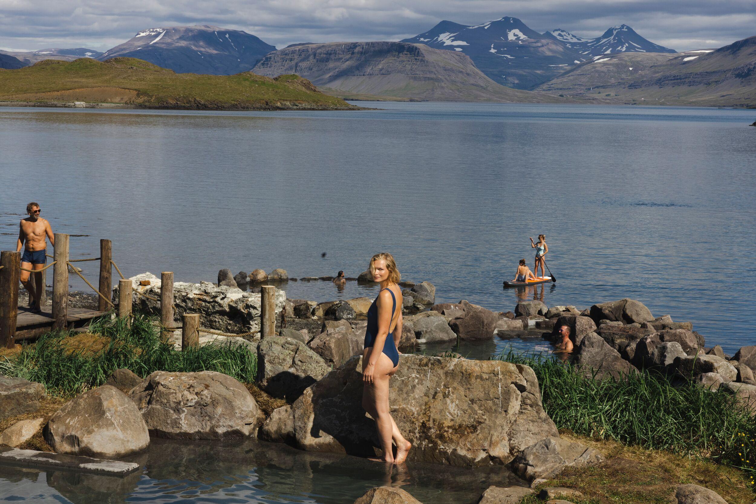 People relax in natural geothermal pools at Hvammsvik Hot Springs in Iceland, with a backdrop of serene water and distant mountains under a partly cloudy sky.