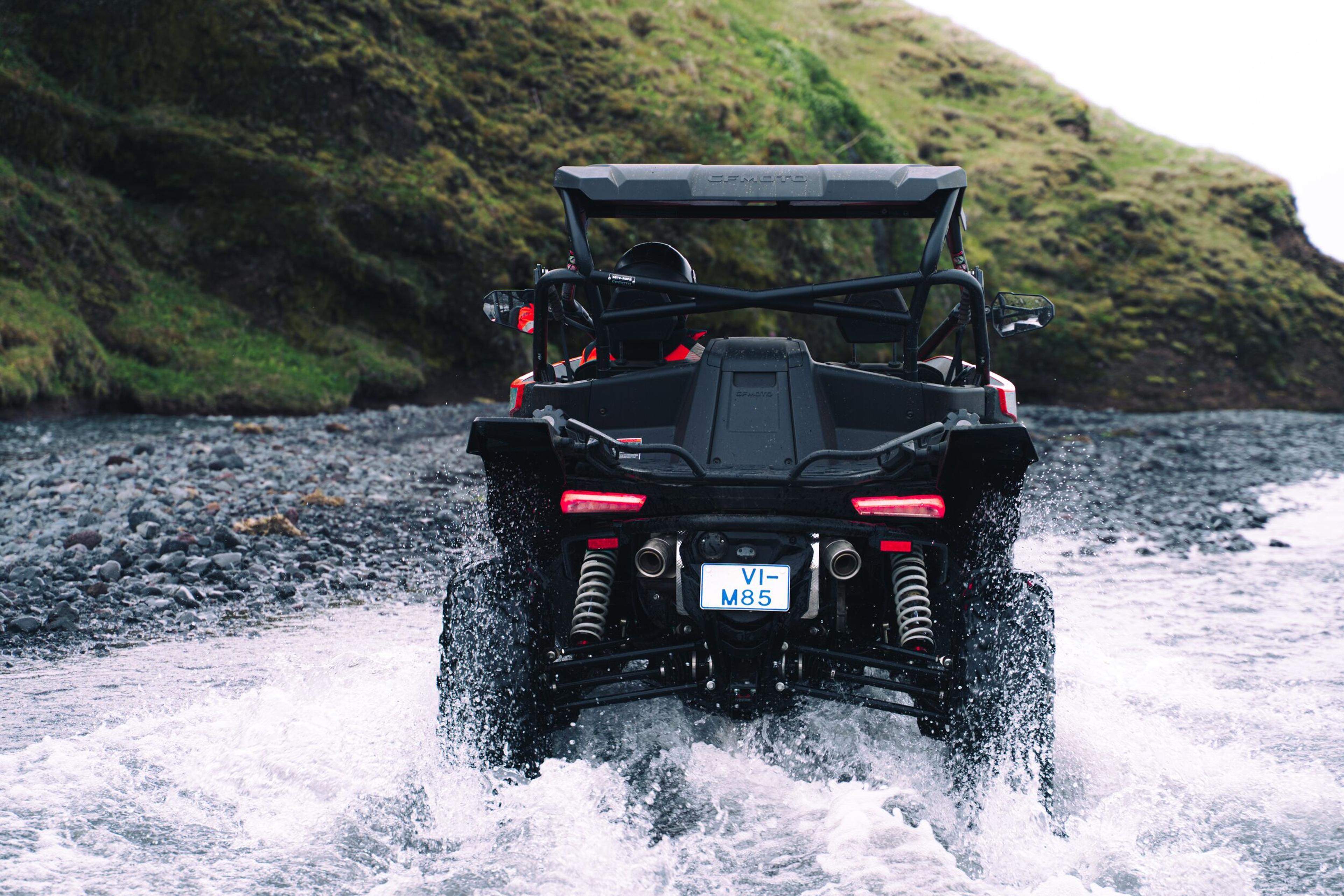 Rear view of a black UTV splashing through water with a green hill in the background.