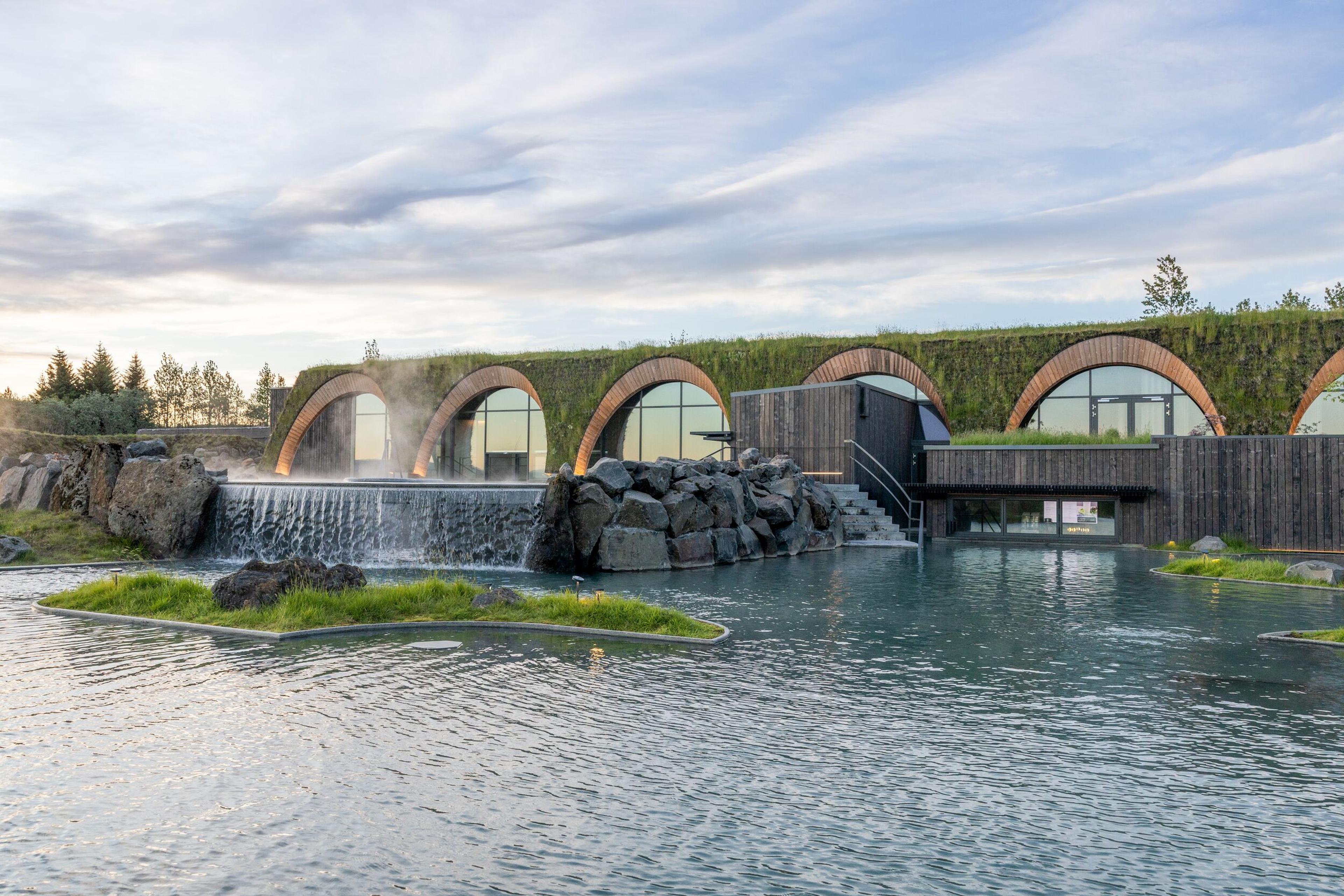 Green-roofed building with arched windows overlooking a turquoise geothermal pool and waterfall.