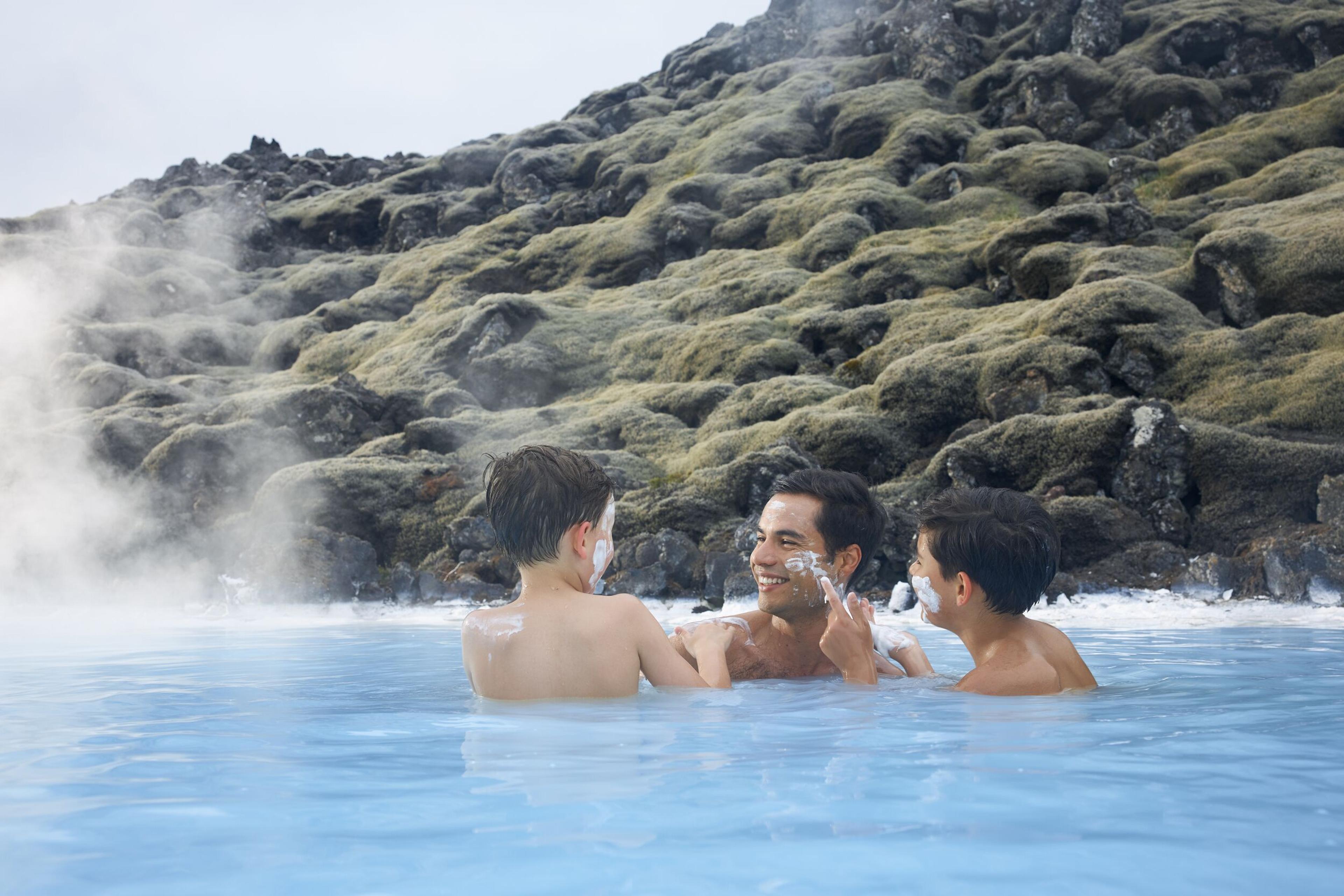 A man and two kids enjoying the milky-blue waters of the Blue Lagoon, playfully applying silica mud masks on each other's faces