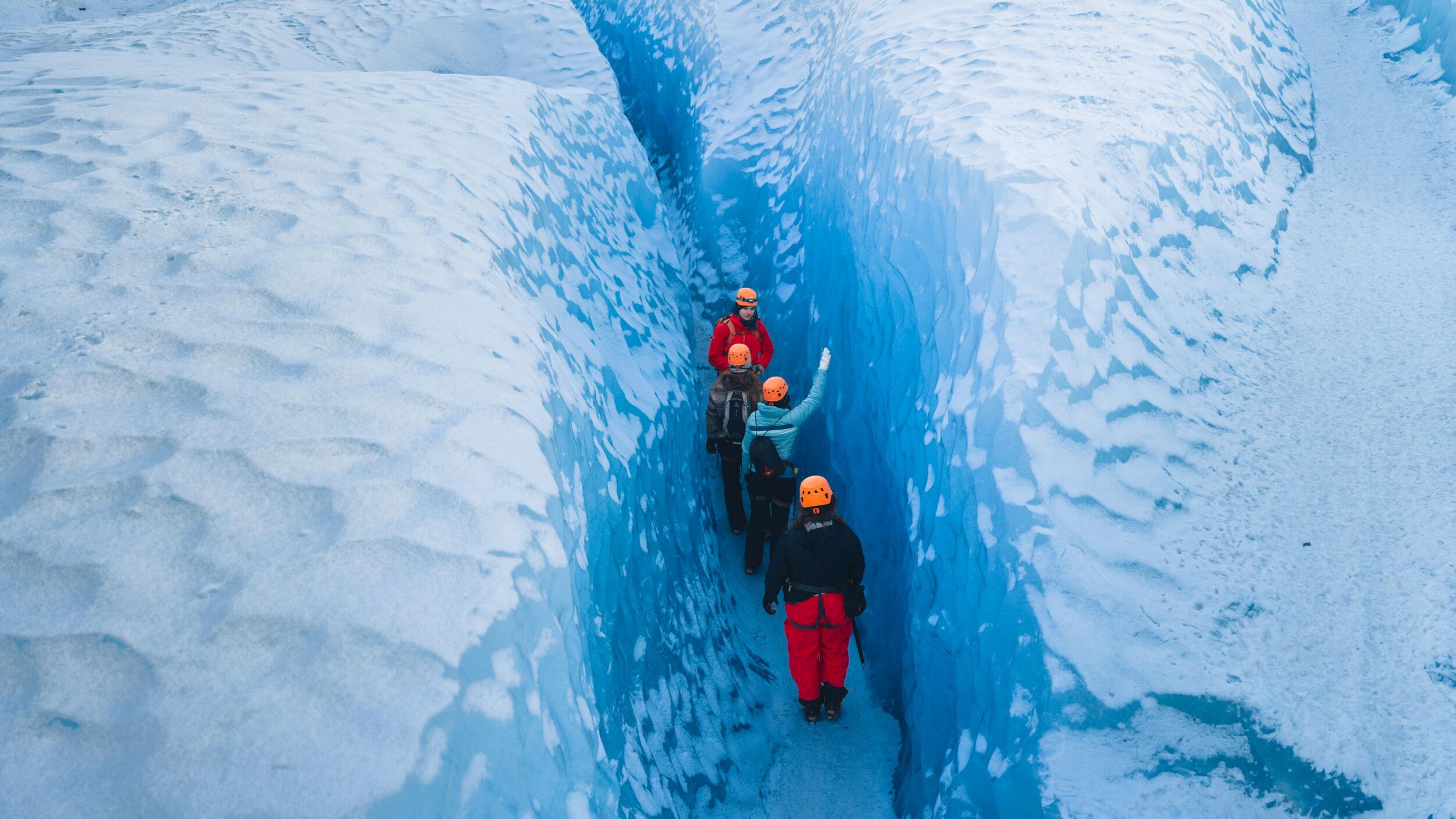 A group of people wearing orange helmets hike through a narrow blue ice crevasse.
