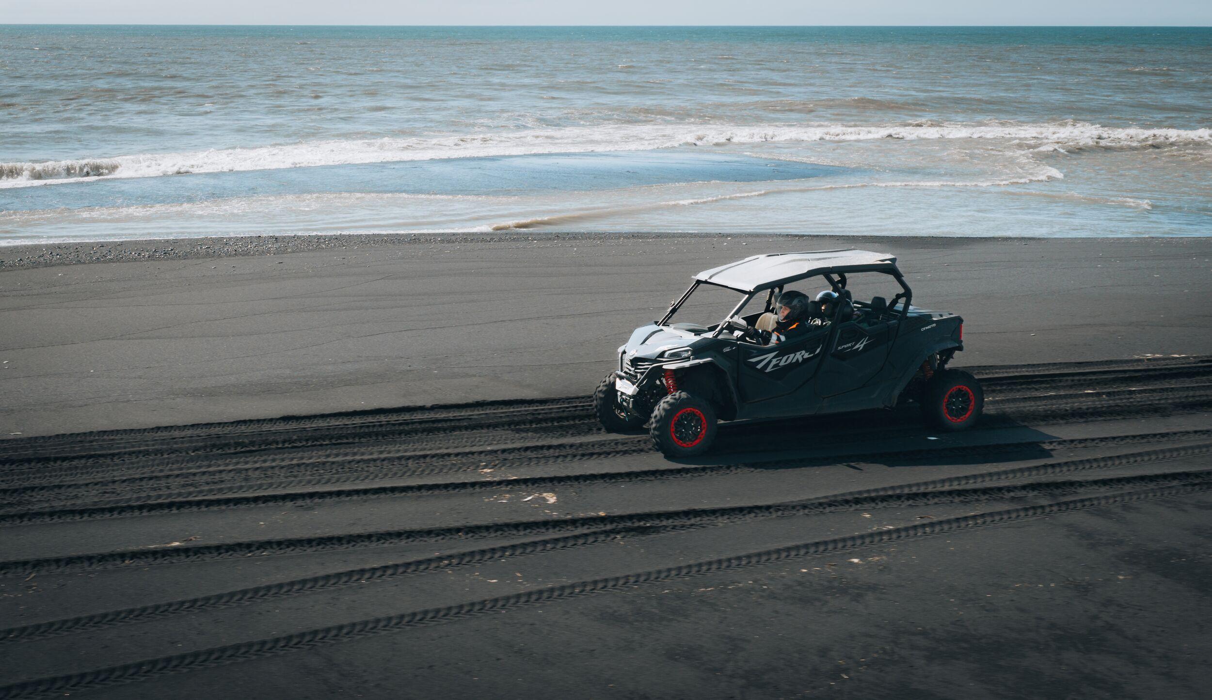 Dark grey side-by-side driving on a black sand beach next to the ocean.