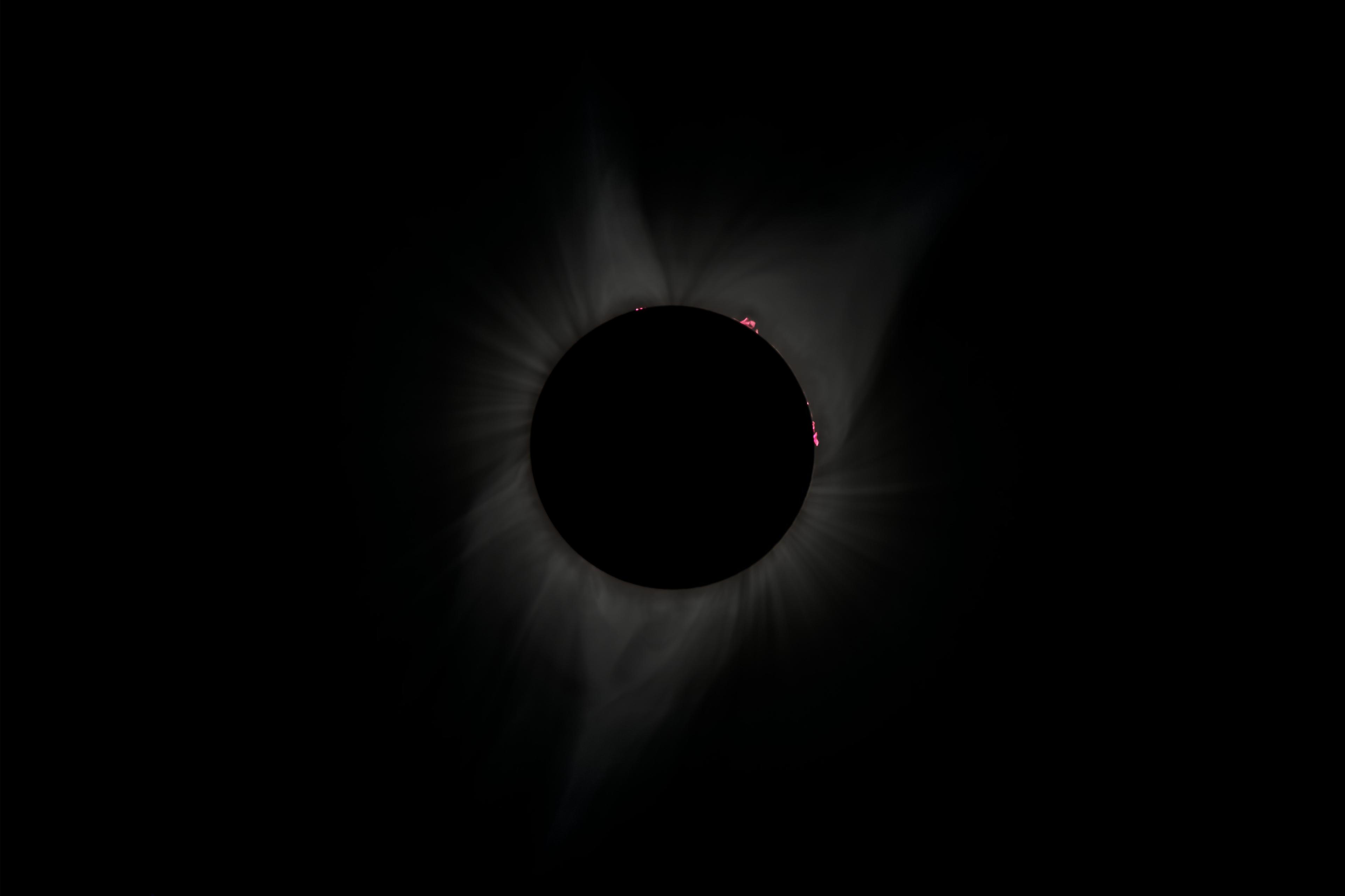 Total solar eclipse with the moon fully blocking the sun, faintly revealing the sun's corona and red solar flares on the edge.