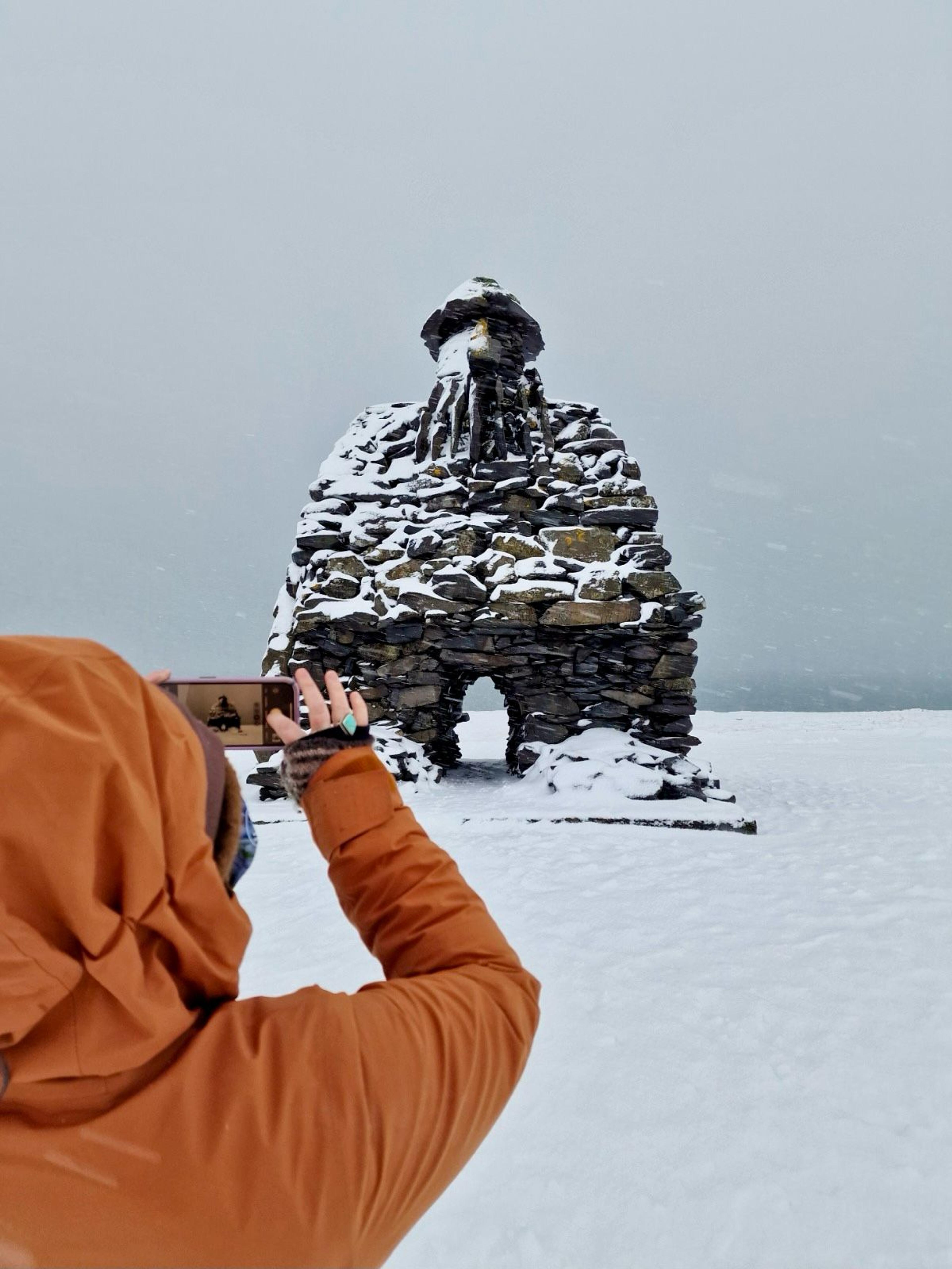 A person in an orange jacket photographs a snow-covered stone cairn with a smartphone.