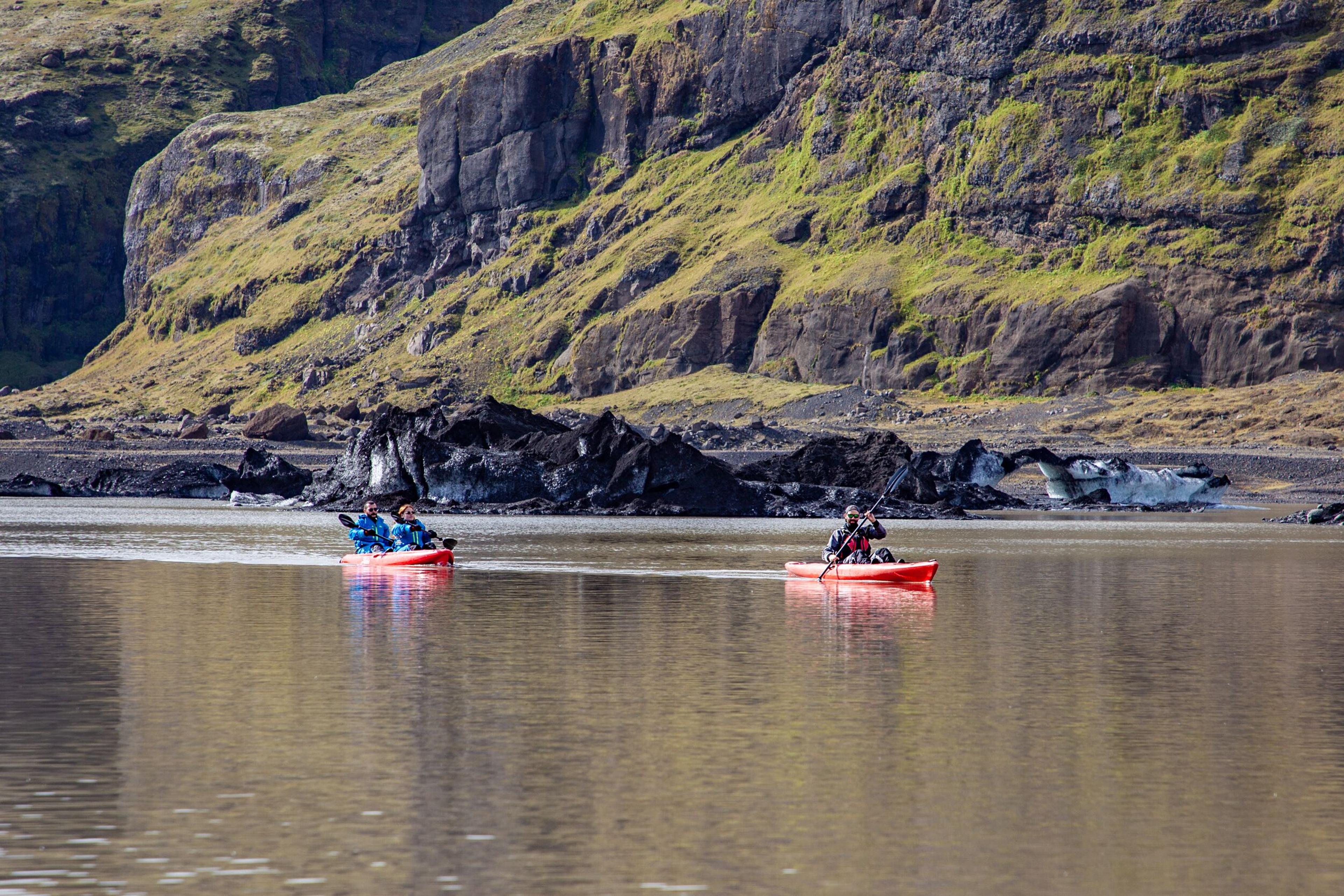 Two red kayaks with kayakers paddle on calm water, with rugged cliffs and rocky terrain in the background, ideal for kayaking in Iceland.