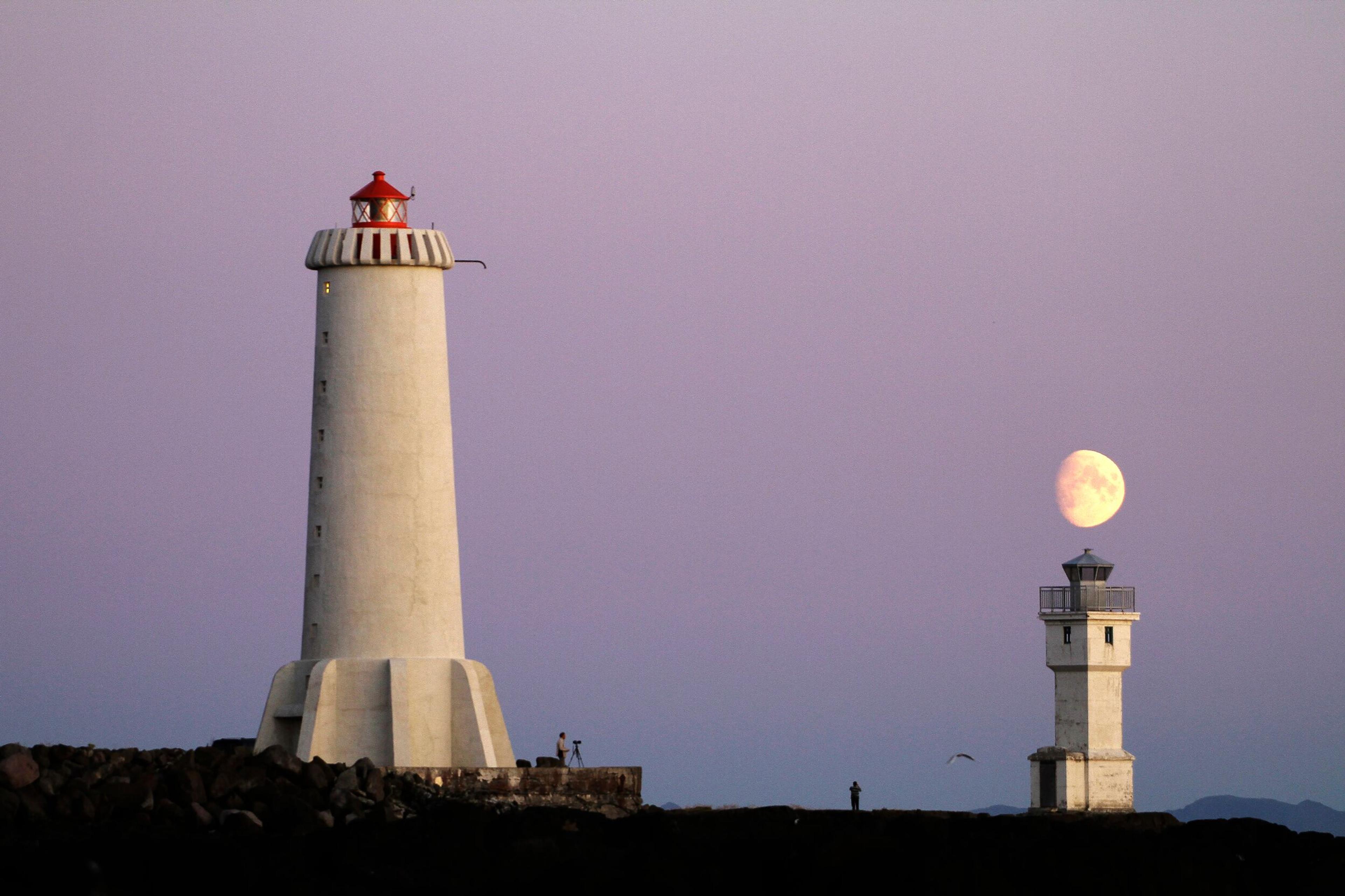 Akranes lighthouse at dusk with a glowing full moon in the background and a smaller lighthouse nearby, set against a serene lavender sky.