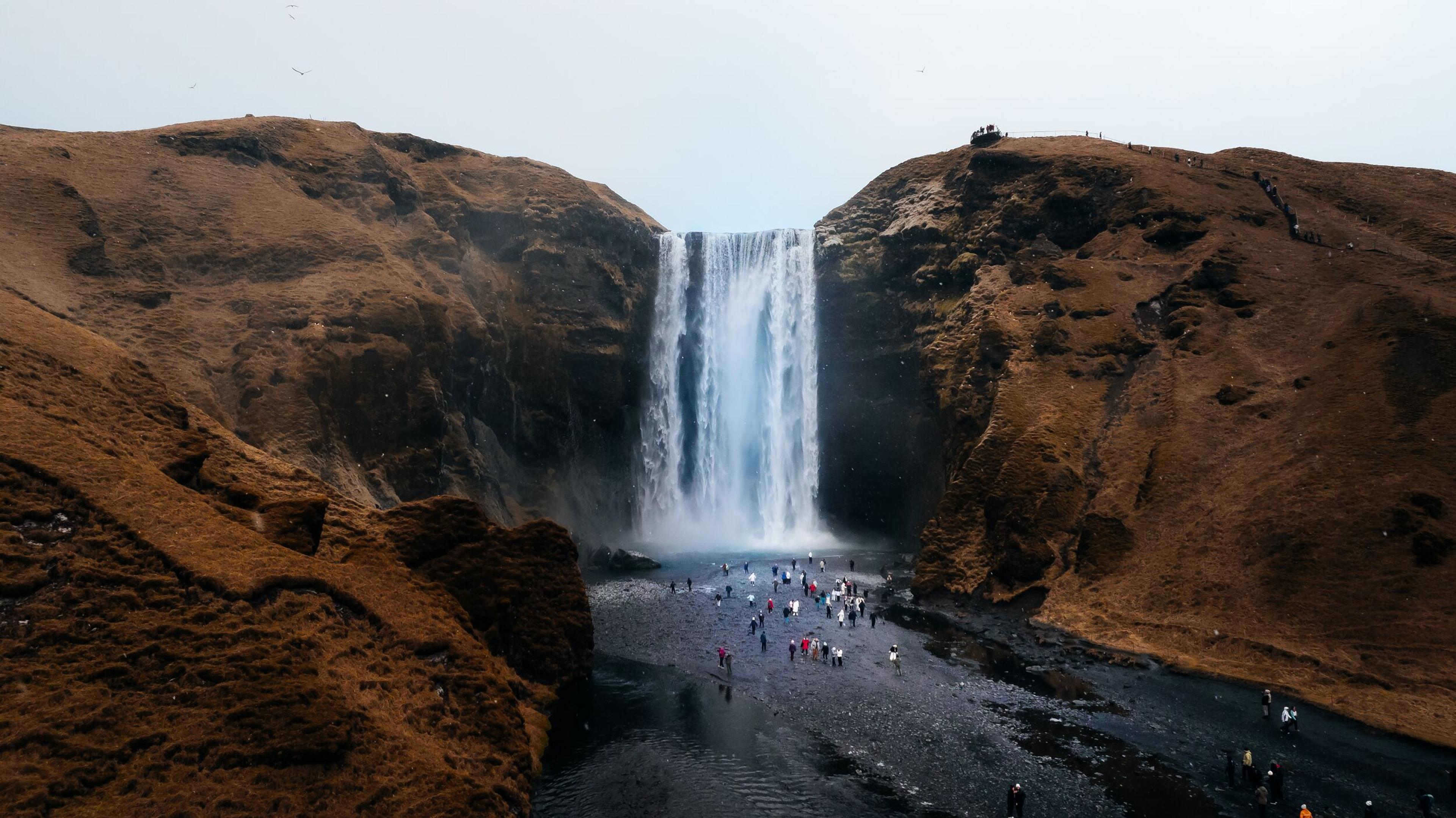 A tall waterfall cascades between two rust-colored hills with many tourists gathered at its base.