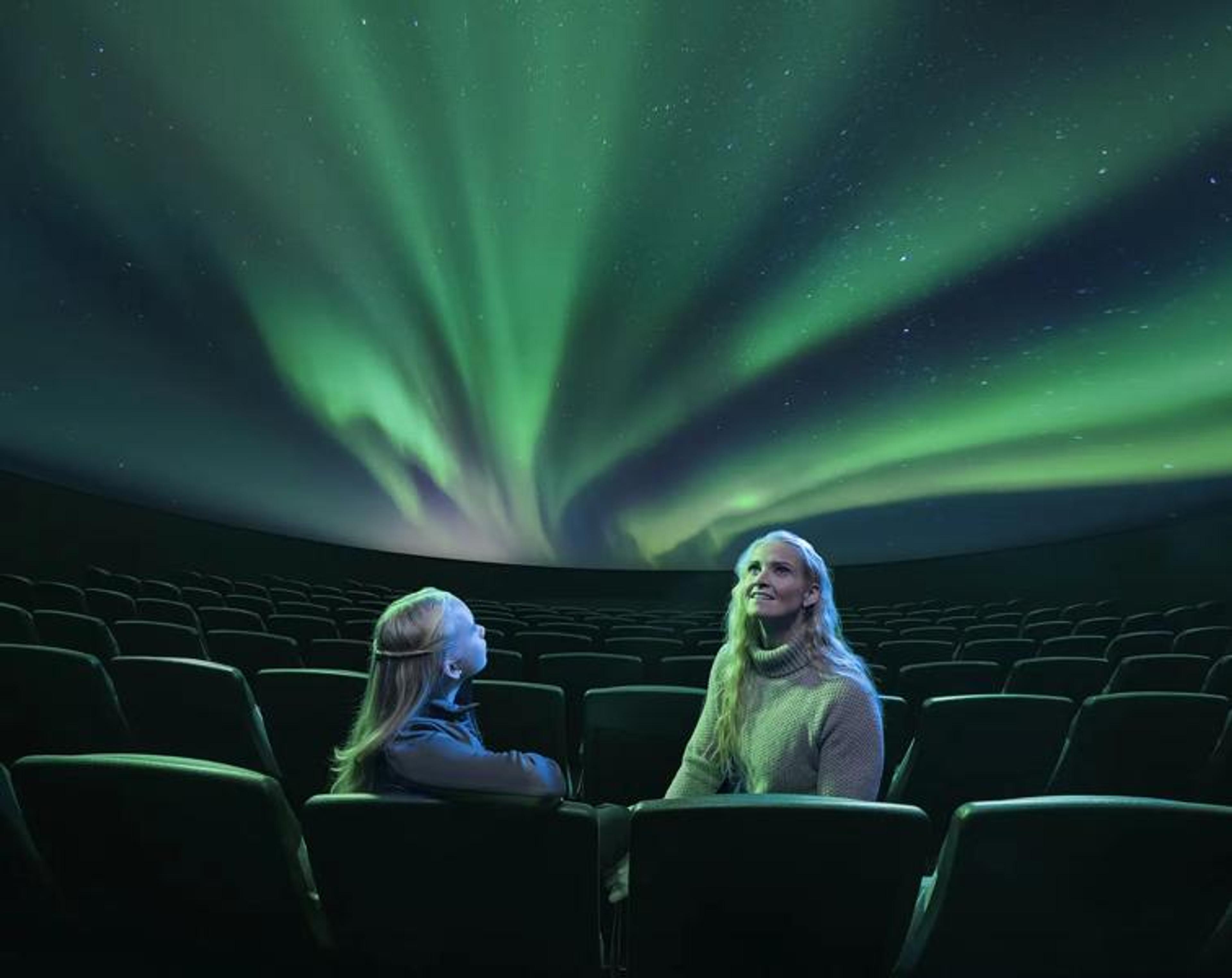 A woman and child in a theater look up at a dome screen displaying the green Northern Lights.
