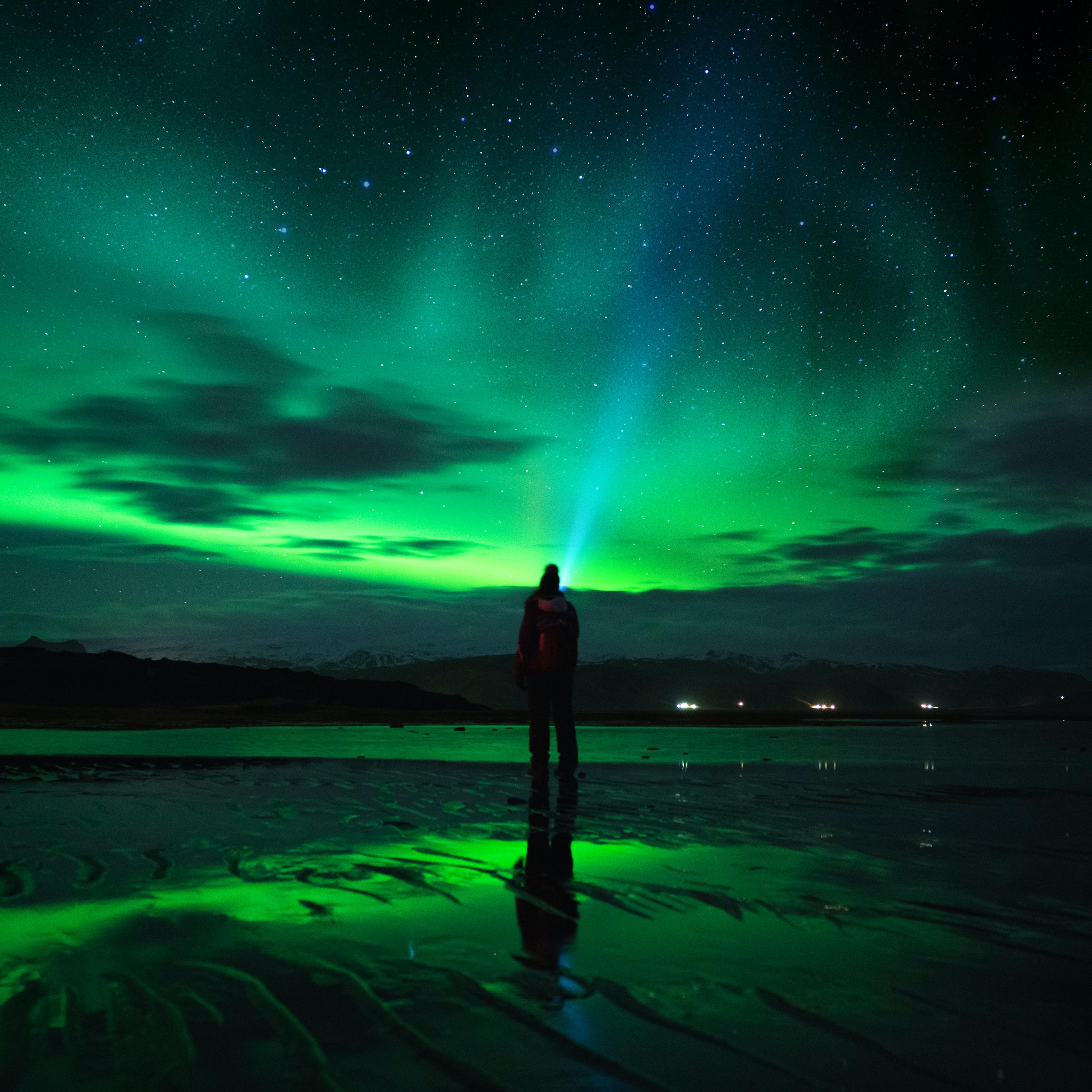 A lone figure stands on a wet beach, backlit by the ethereal glow of the Northern Lights and a starry sky.