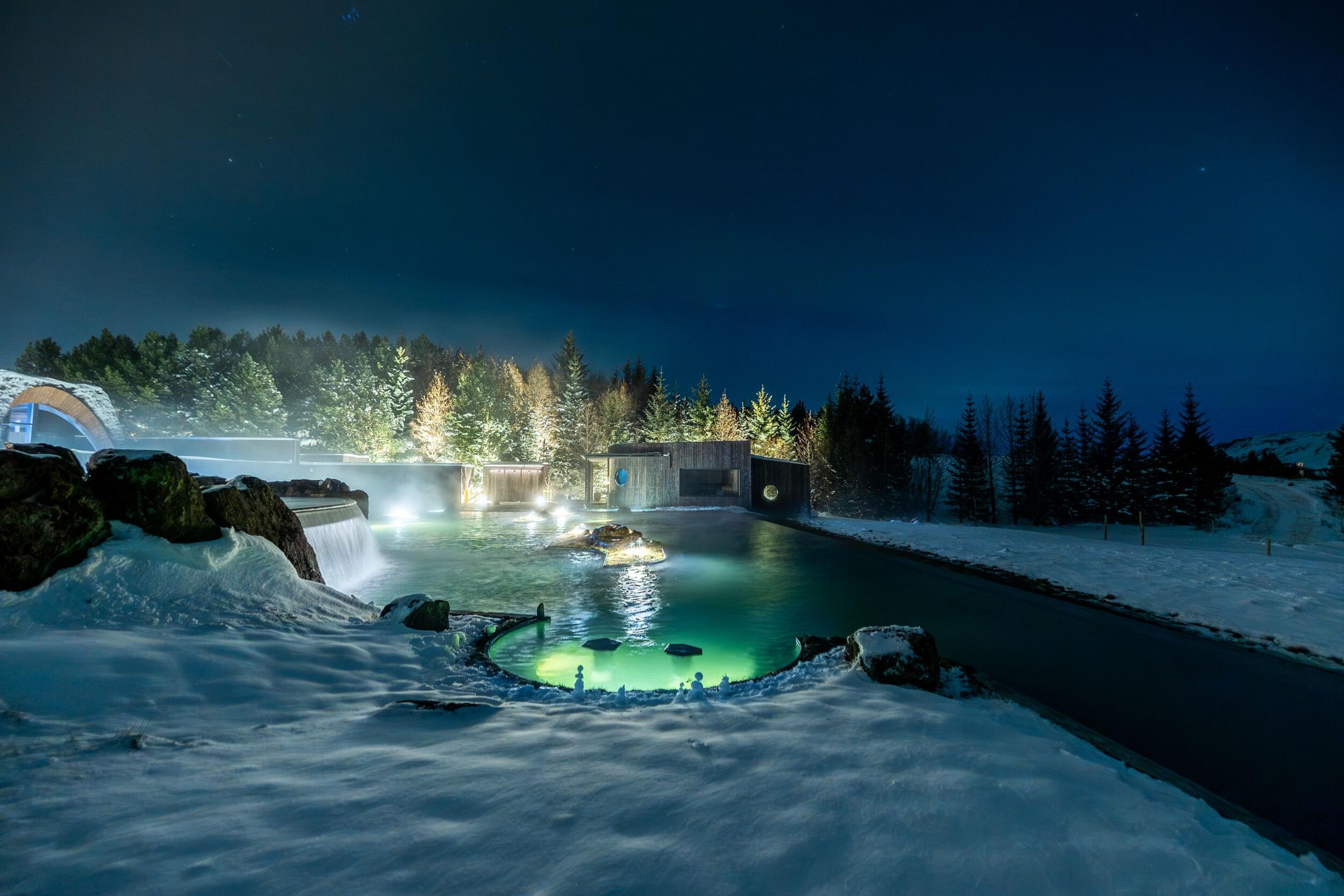Snowy landscape at night with illuminated blue-green geothermal lagoons, a waterfall, and lit evergreen trees.