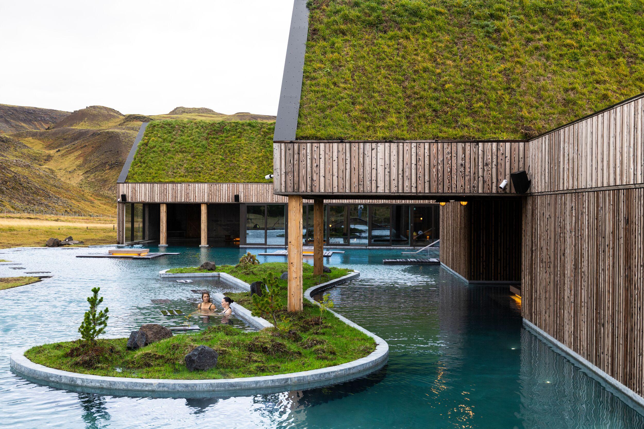 Two people relaxing in a geothermal pool next to modern wooden buildings with grass-covered roofs, in a hilly landscape.