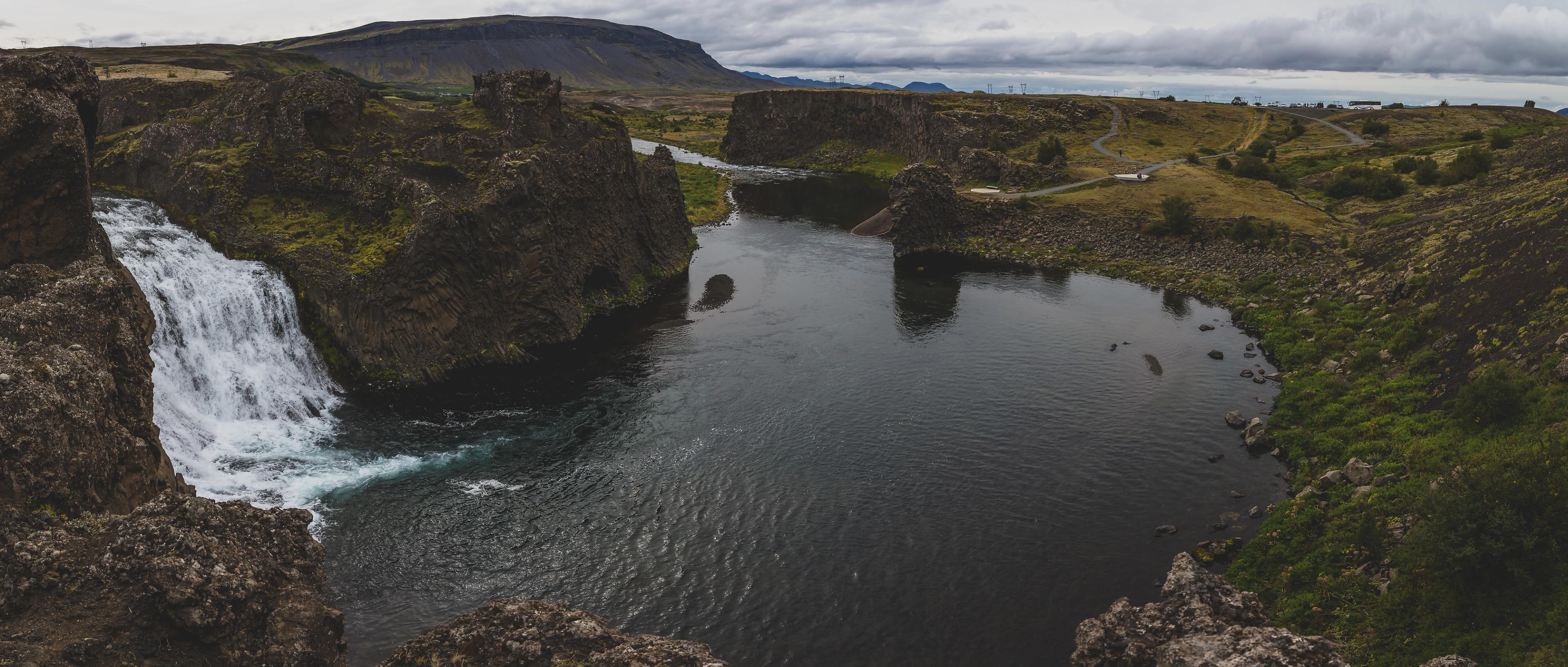 A powerful waterfall plunges into a dark river surrounded by rugged, mossy volcanic rock and distant hills under a cloudy sky.