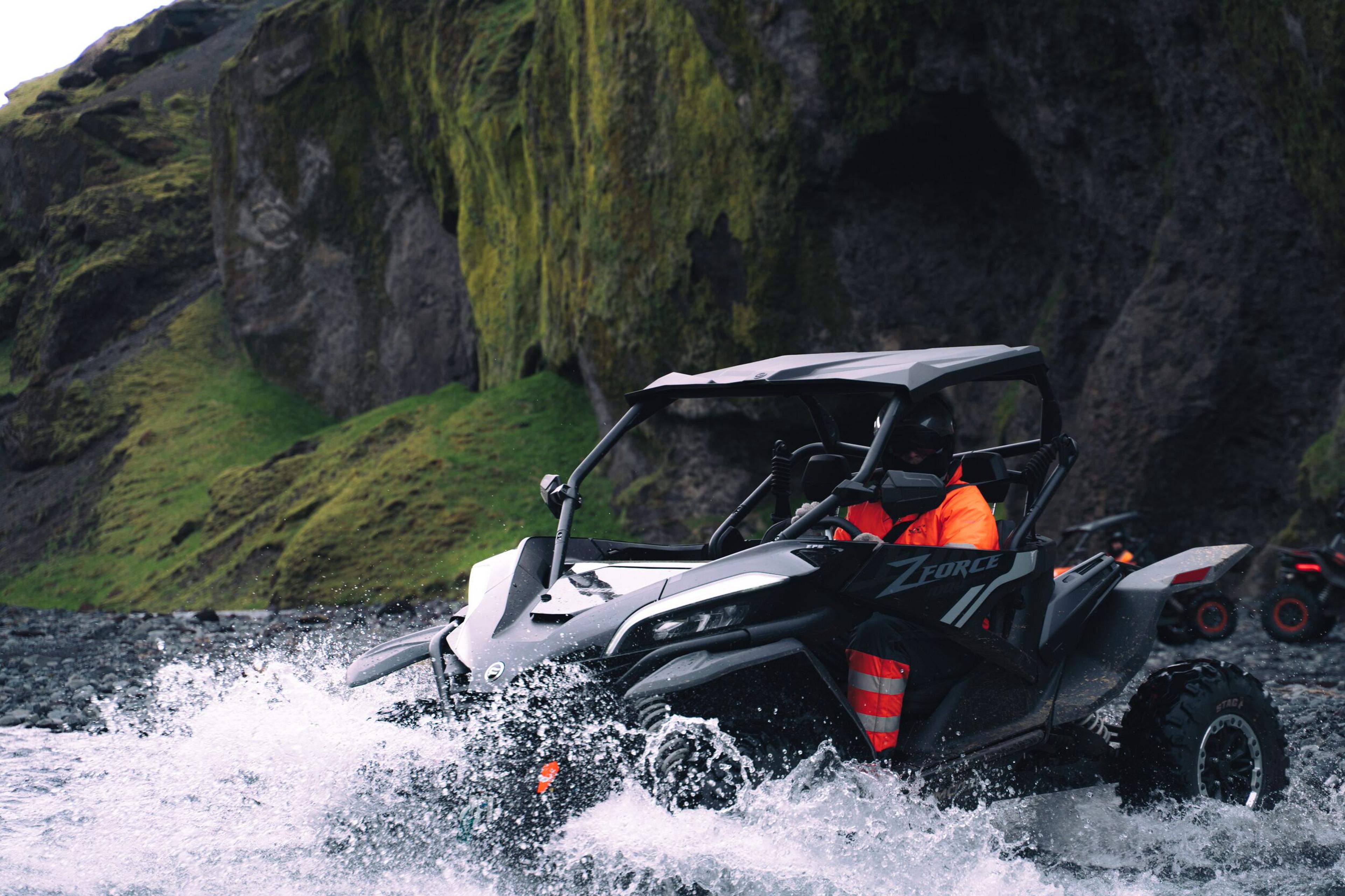 A black UTV with a driver in orange splashes through a shallow river, with green mossy cliffs in the background.