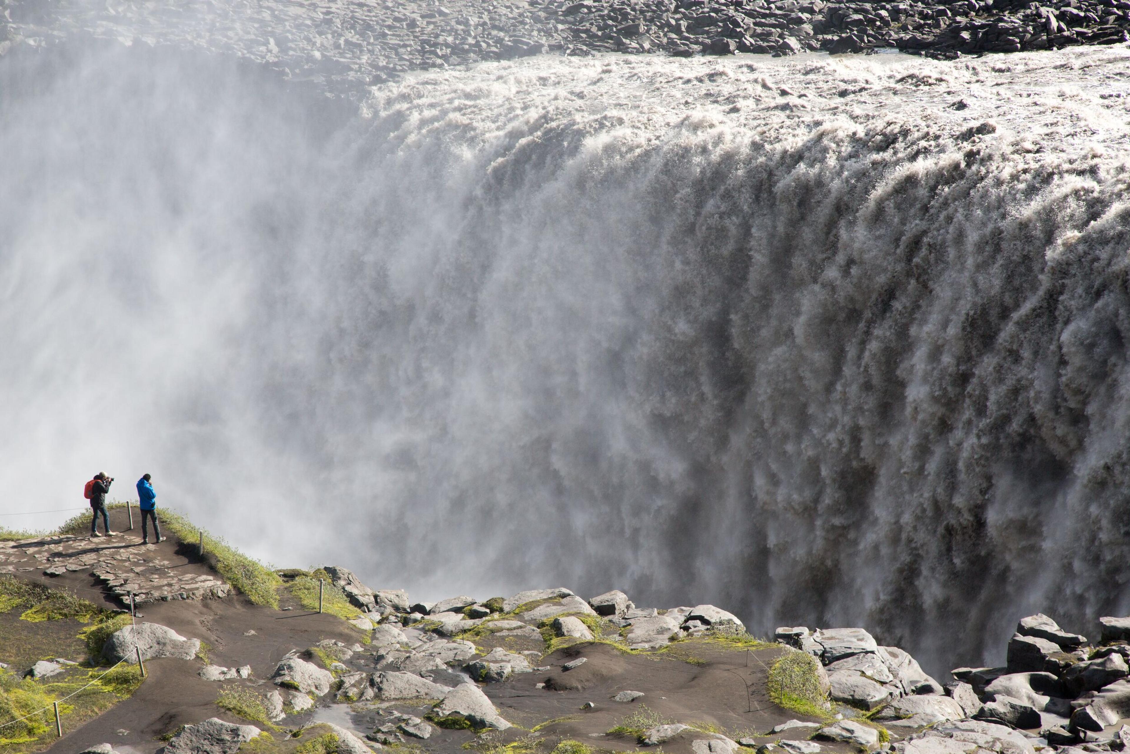 Two individuals capturing photos of Dettifoss, recognized as the most powerful waterfall in Iceland.