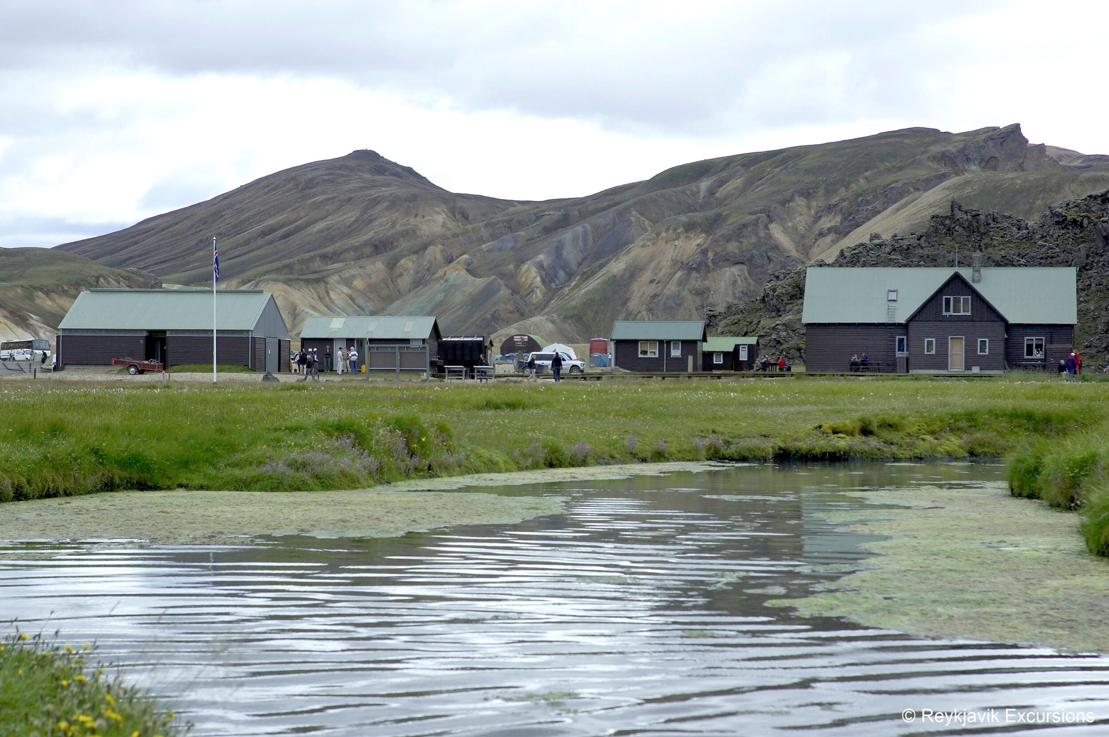 A pond in a grassy field fronts several dark buildings, one with an Icelandic flag, backed by colorful, rugged mountains under a cloudy sky.
