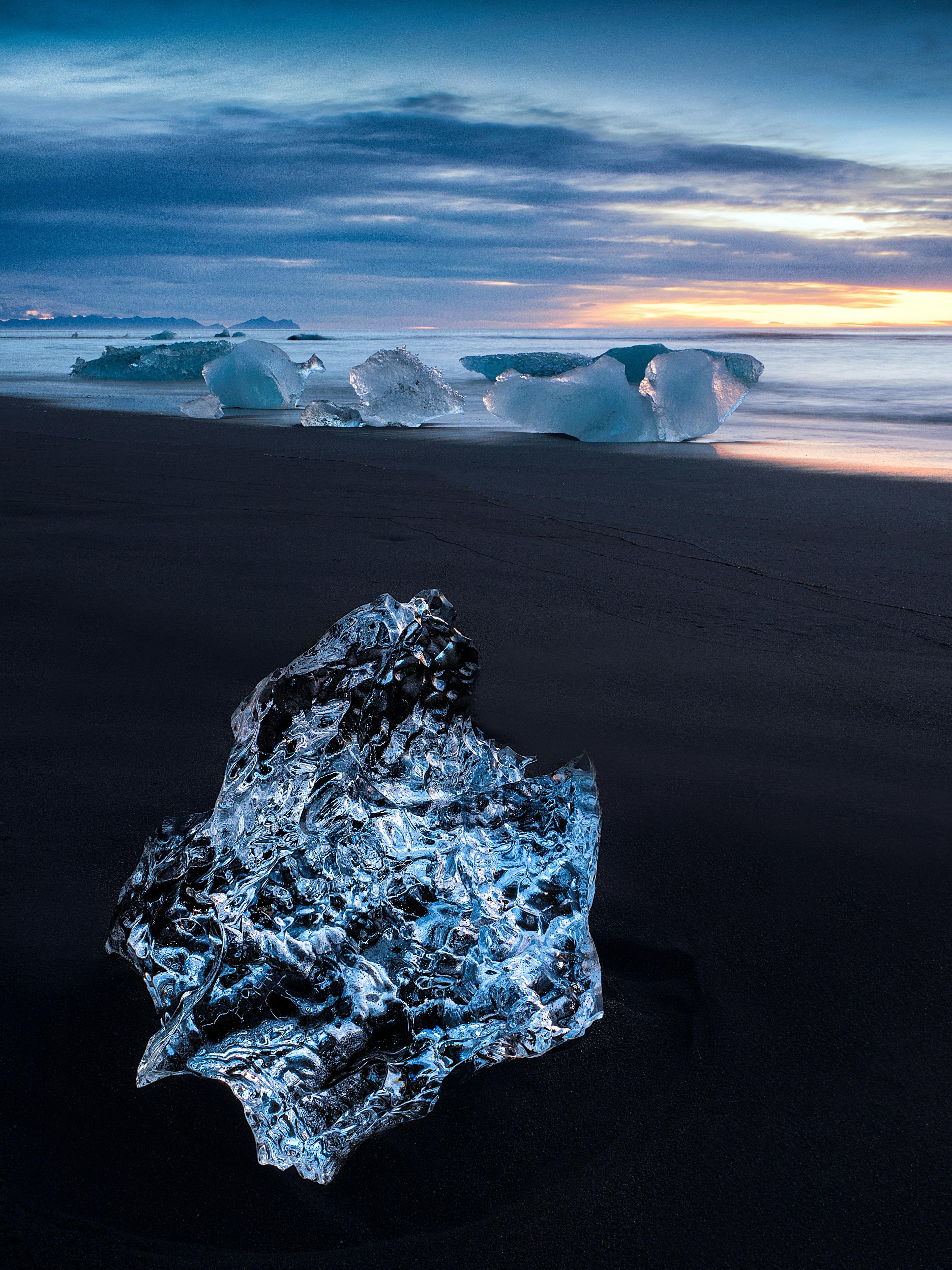 A piece of clear glacial ice on a black sand beach with more icebergs in the background and a colorful twilight sky reflected on the wet sand.