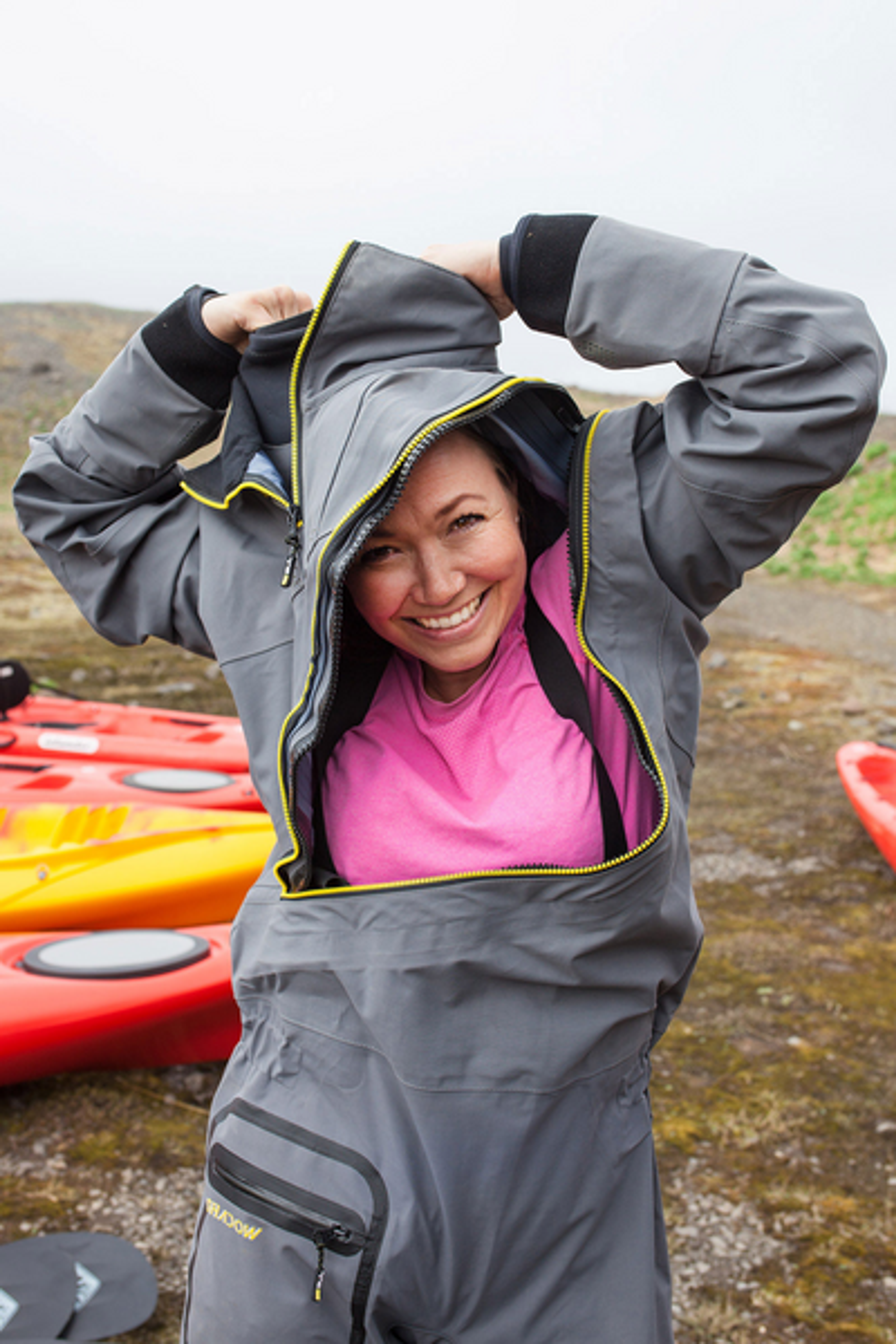 A smiling woman in a pink shirt puts on a gray dry suit with red kayaks in the background, ready for kayaking in Iceland.