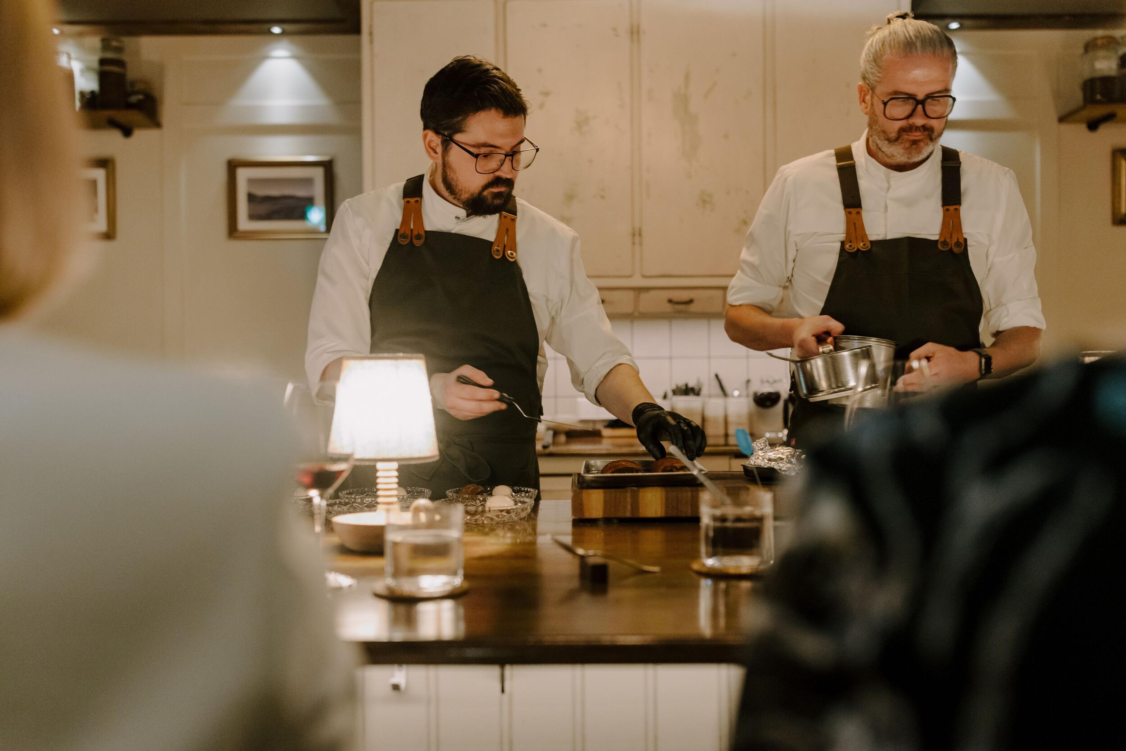 Two chefs in aprons prepare food behind a counter, with a blurred diner in the foreground.