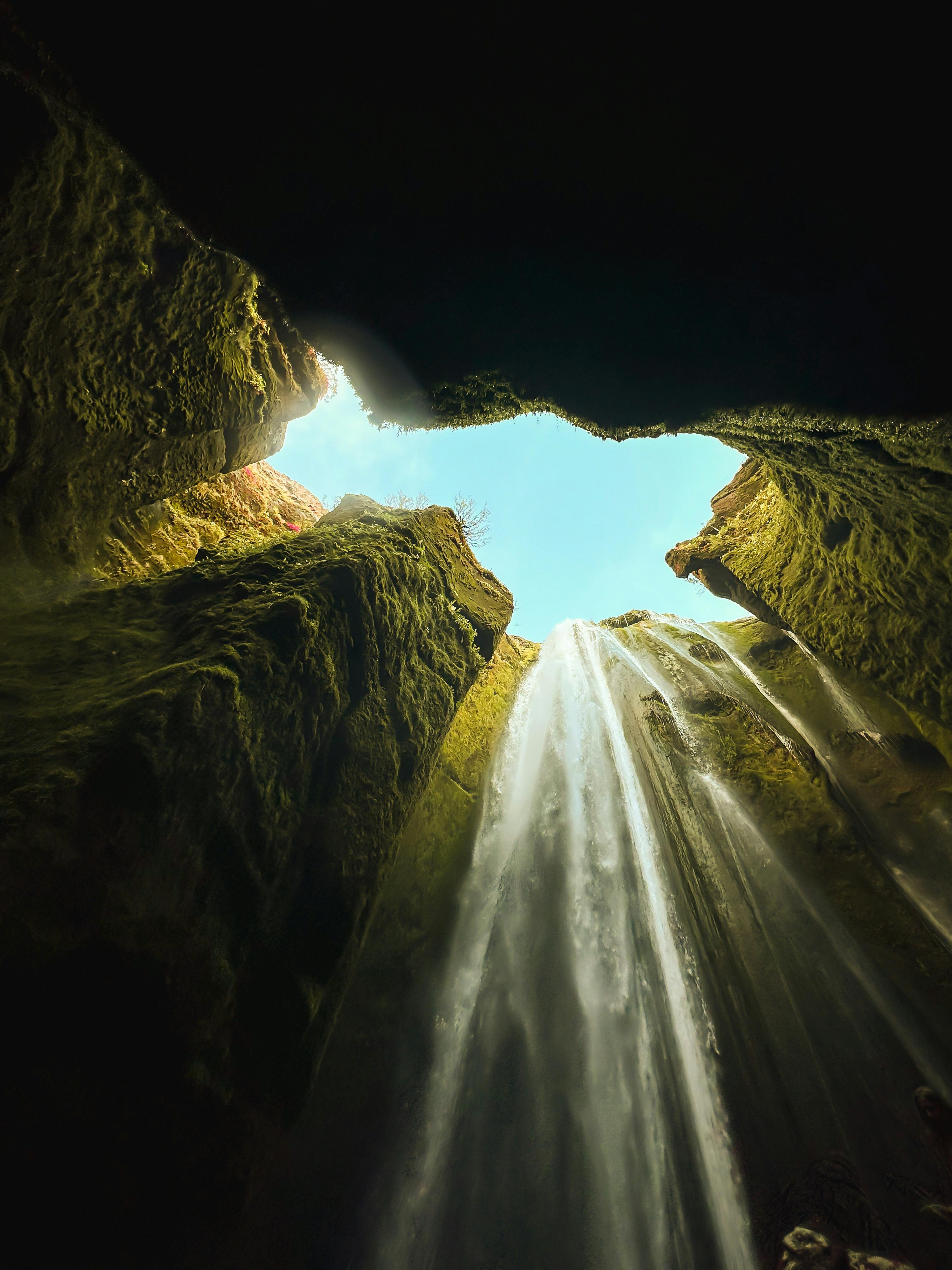 Looking up from the base of Gljúfrabúi waterfall, with water streaming down moss-covered cliffs and a patch of blue sky visible above.