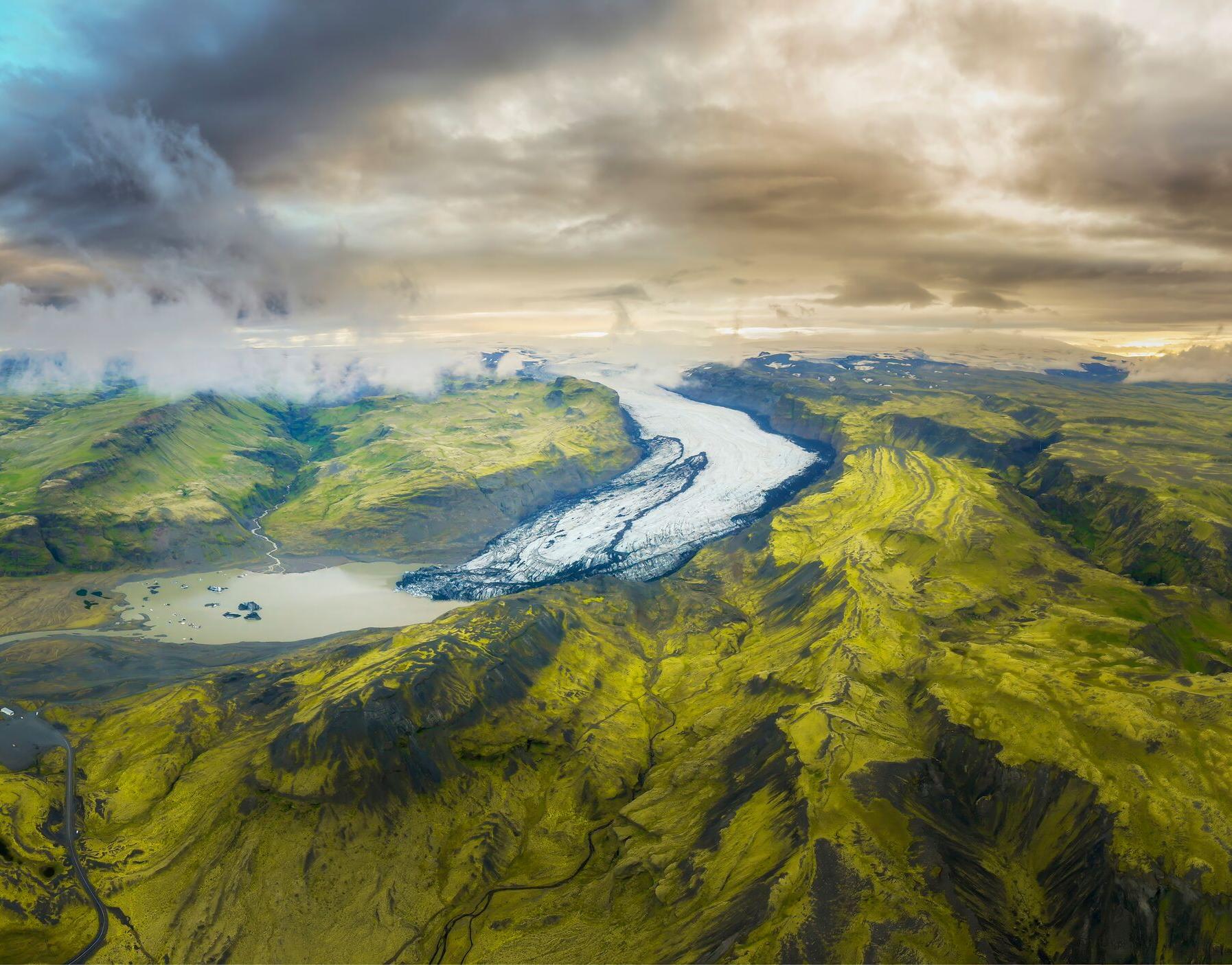 Aerial view of a winding glacier flowing through vibrant green landscapes under a dramatic sky in Iceland, illustrating the stunning natural beauty of the region.