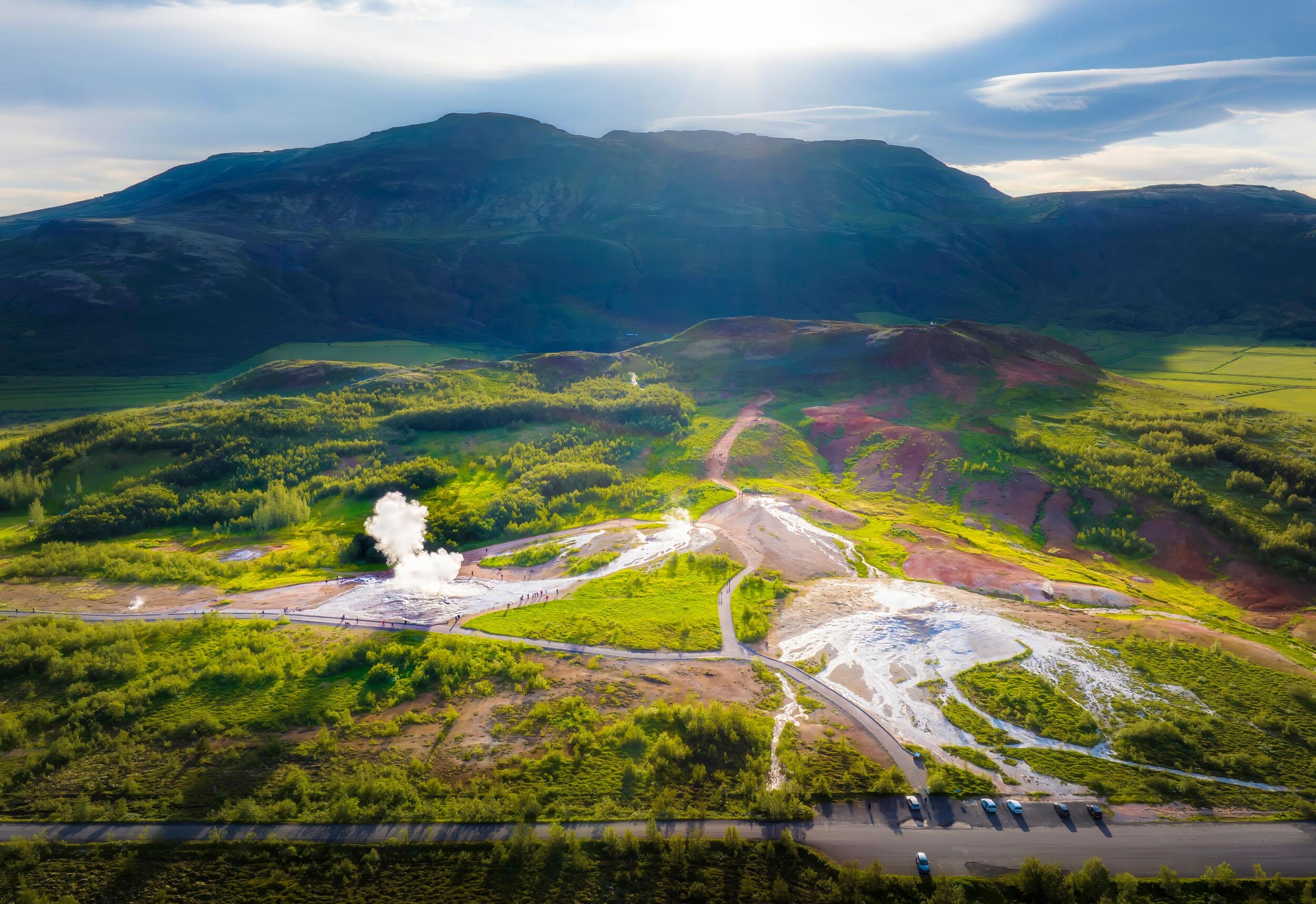 Aerial view of the geothermal area in Iceland, with a geyser erupting, casting a plume of steam against the lush green landscape and rugged mountains in the background, under the soft glow of sunlight.