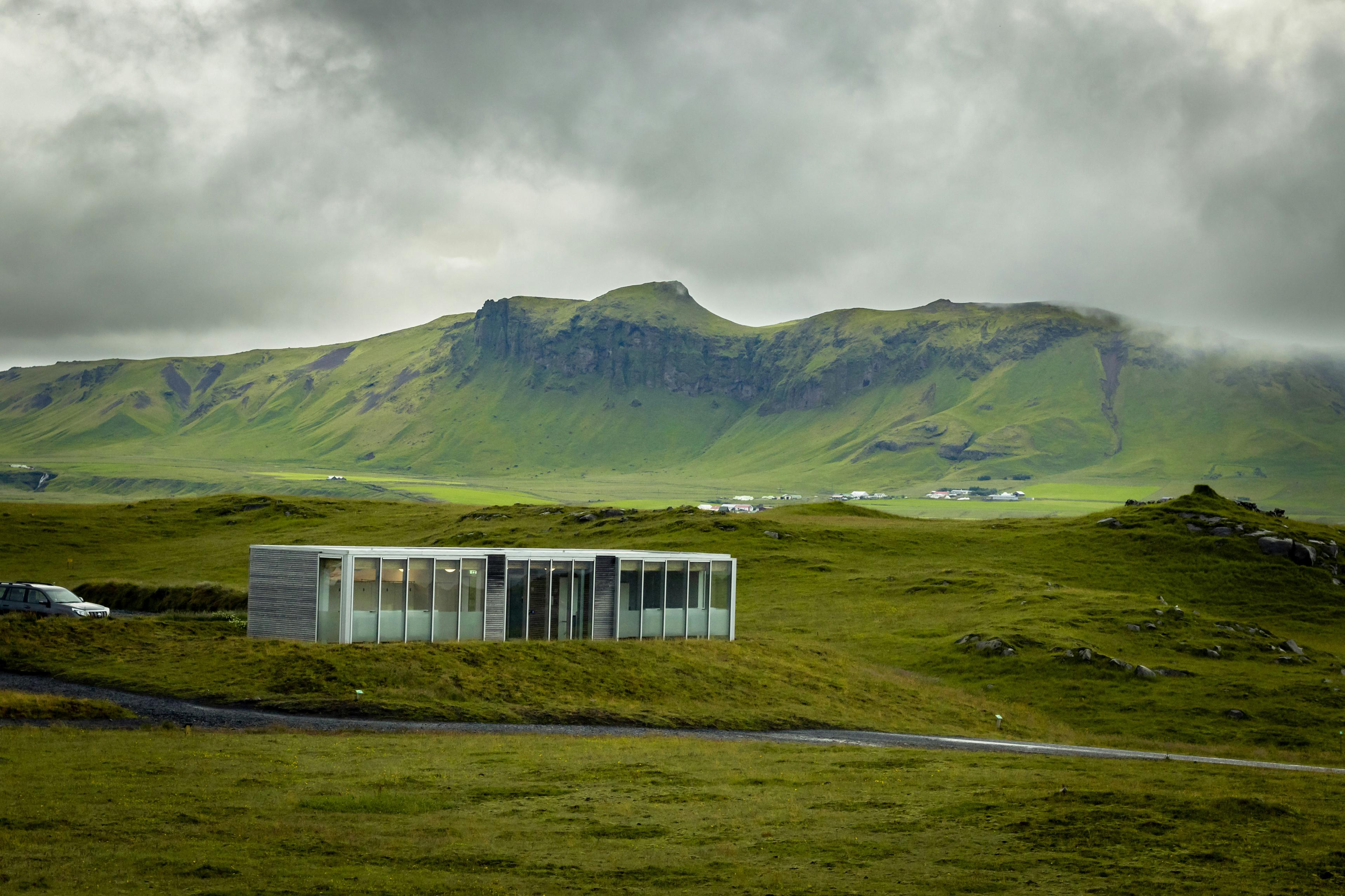 Modern glass-fronted cabin in a green, mountainous landscape near Vík, Iceland, under a cloudy sky.