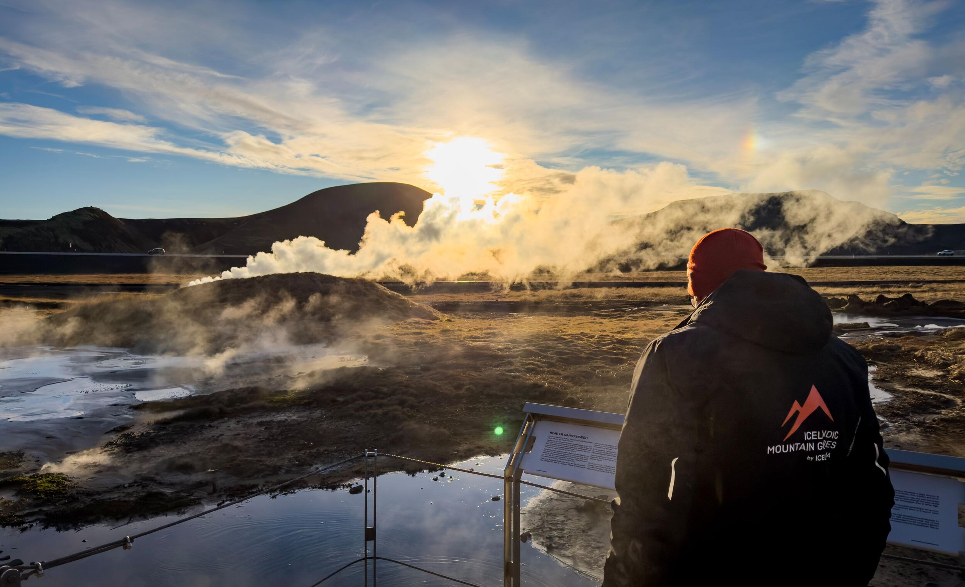 Person in a red beanie looks at a steaming geothermal field with hot springs under a bright, low sun.