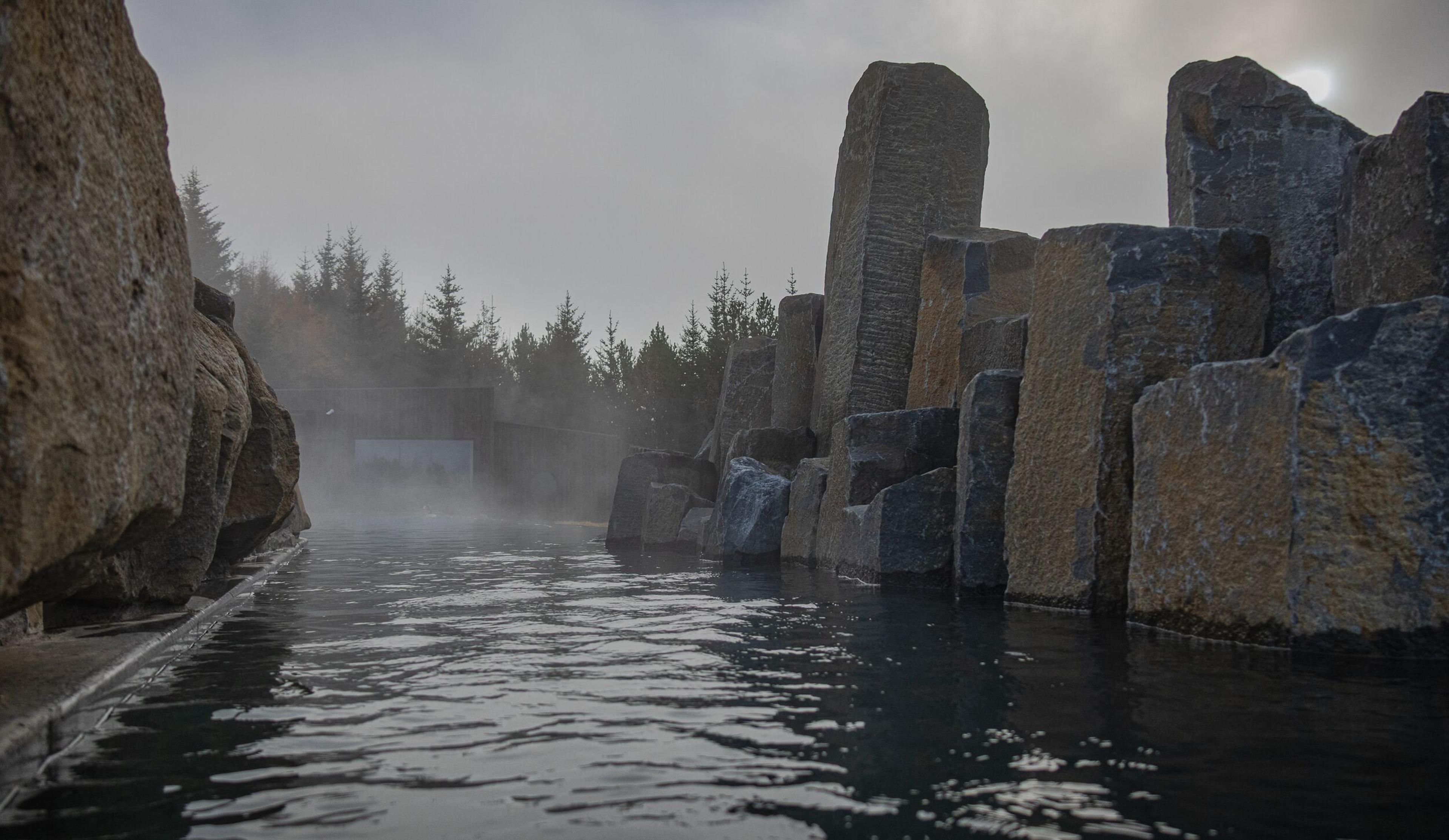 Steaming dark water surrounded by tall, geometric rock columns and a misty forest.
