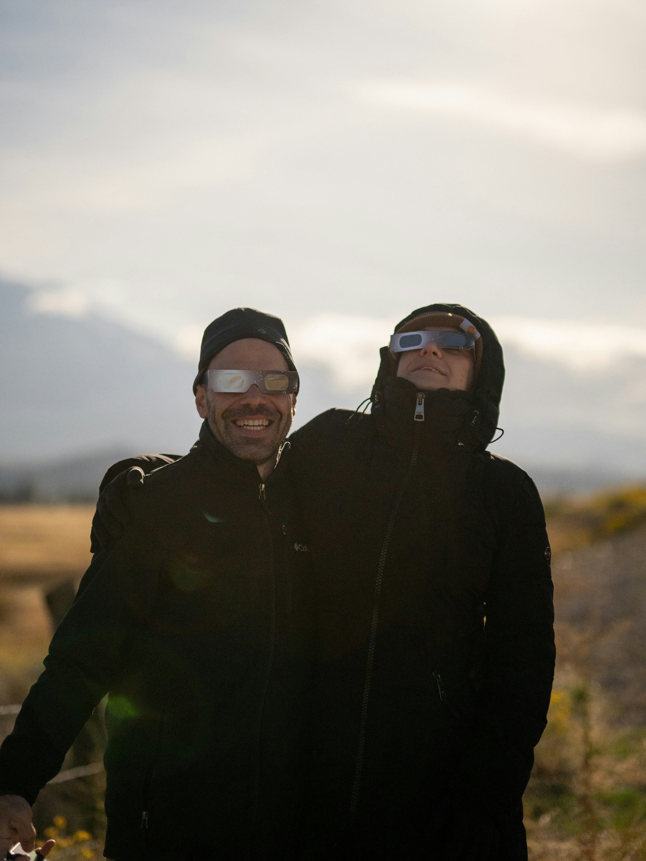 Two people wearing protective glasses, smiling while observing a solar eclipse, dressed in dark jackets outdoors under dim lighting.