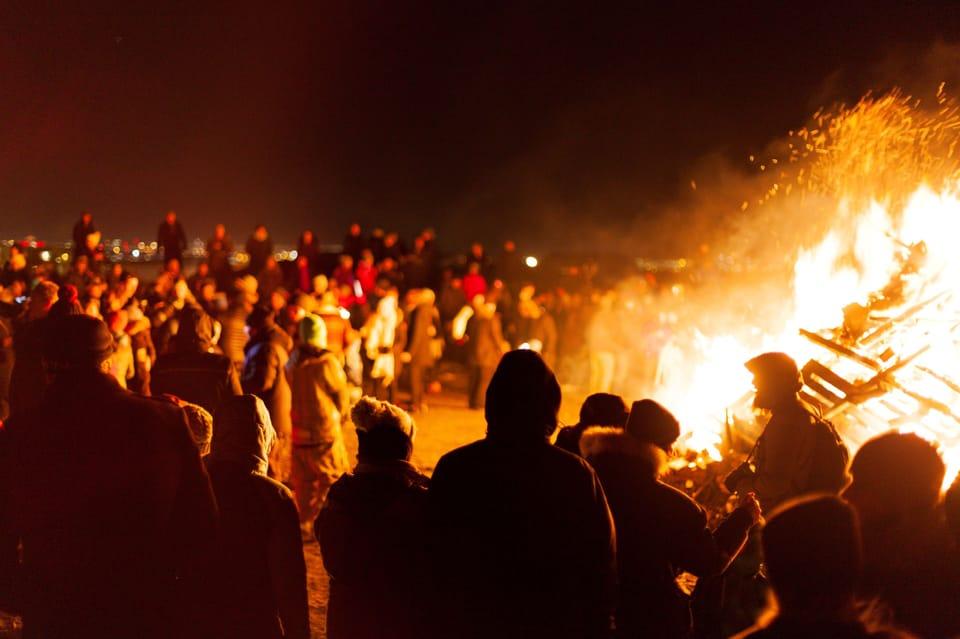 Crowd gathered around a large bonfire during Reykjavik's New Year's Eve celebration, with people warmly dressed and illuminated by the fire's glow. The night sky and distant city lights add to the festive atmosphere as people enjoy the traditional Icelandic bonfire event.