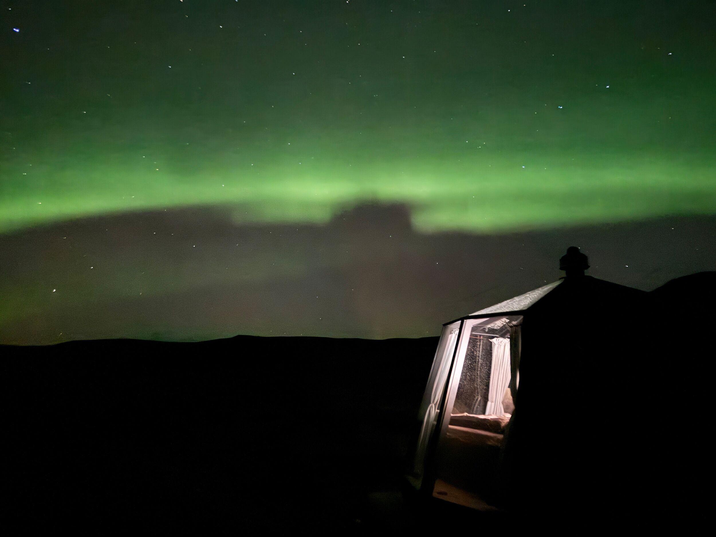 A modern tent with a lit interior under a bright green aurora borealis and starry night sky.