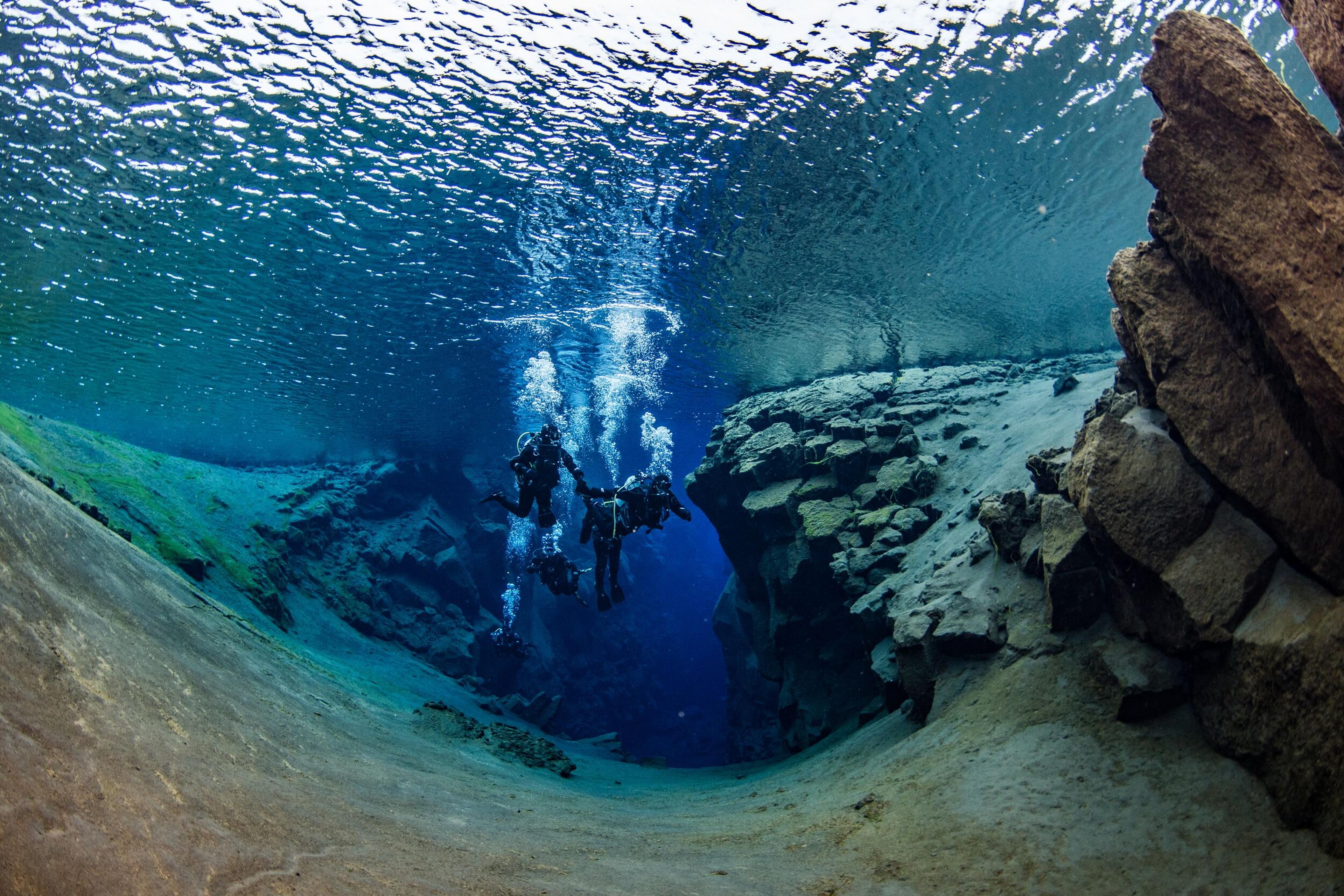 Scuba divers explore a clear blue underwater fissure with rocky walls.