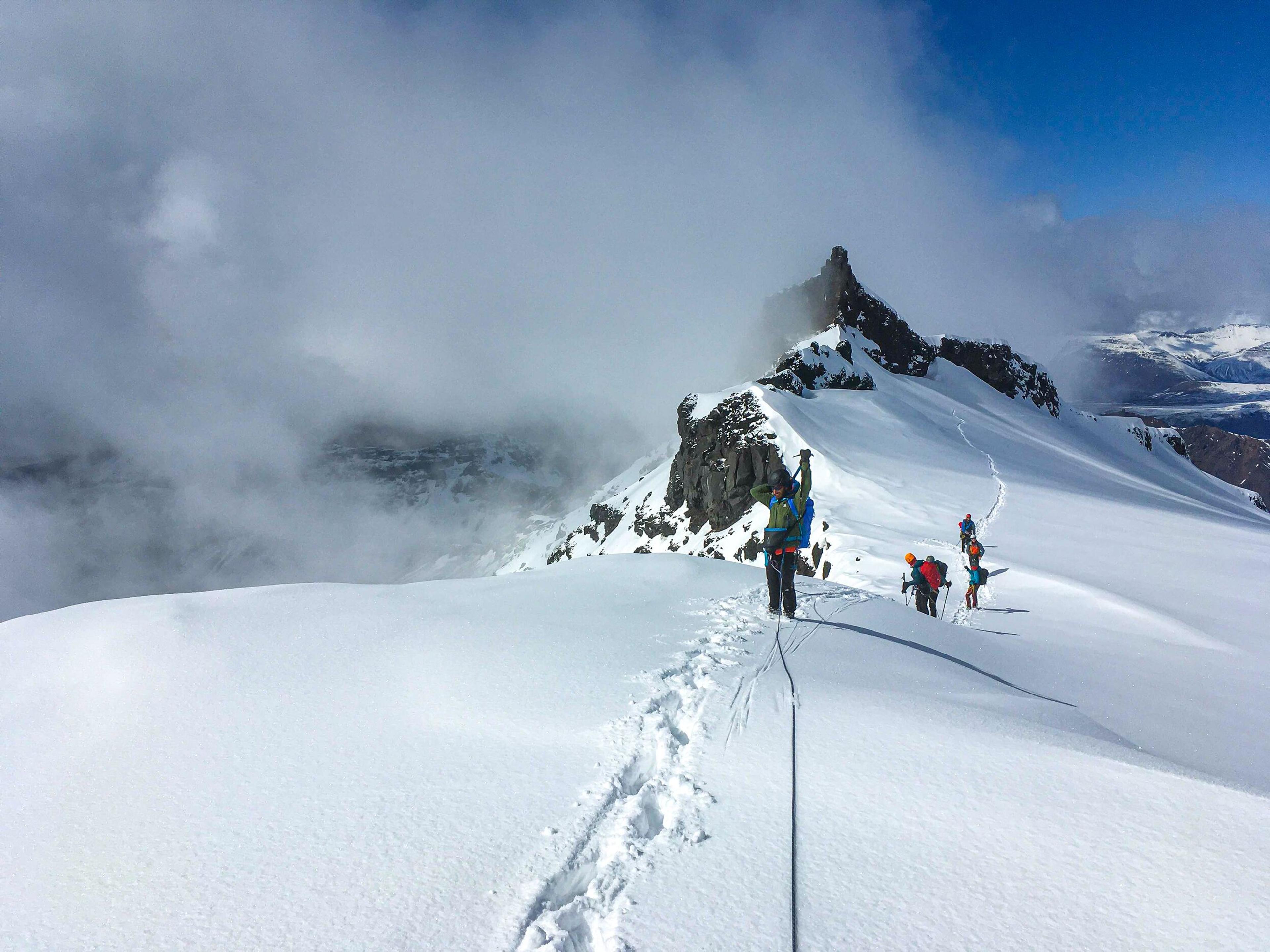 Mountaineers ascend a snow-covered ridge, roped together for safety, with a rocky peak piercing the clouds in the background.