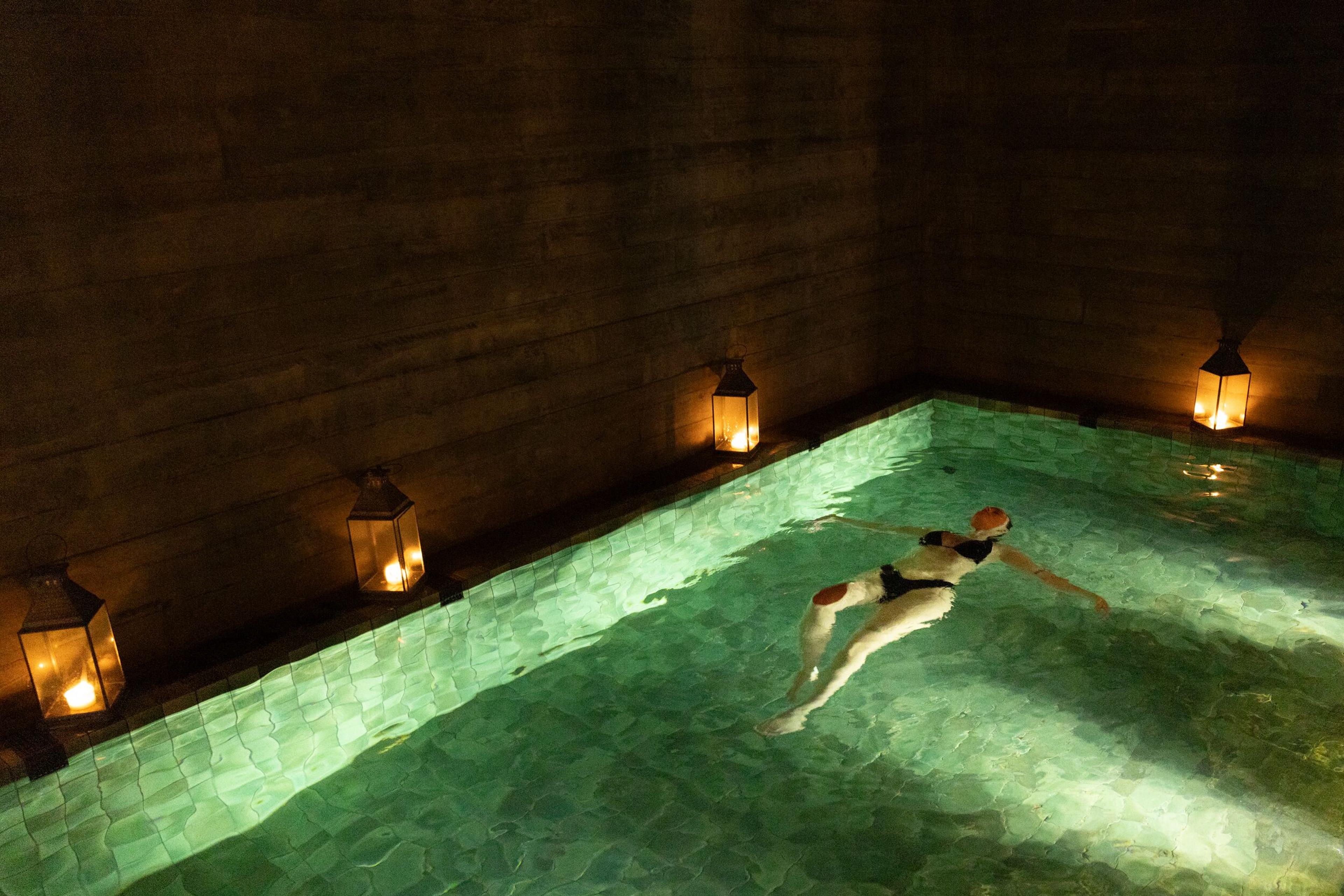 A person floats on their back in a dimly lit pool, surrounded by lanterns casting a warm glow.