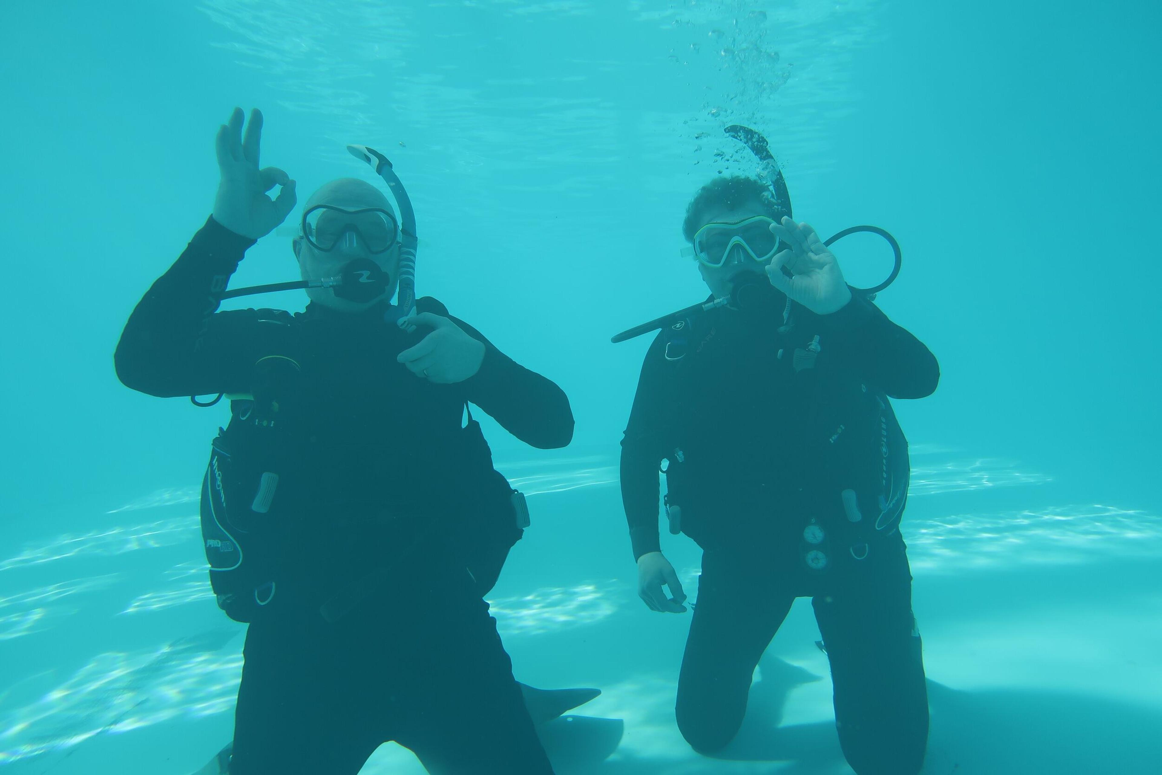 Two scuba divers underwater in a pool, both giving the "OK" hand signal.