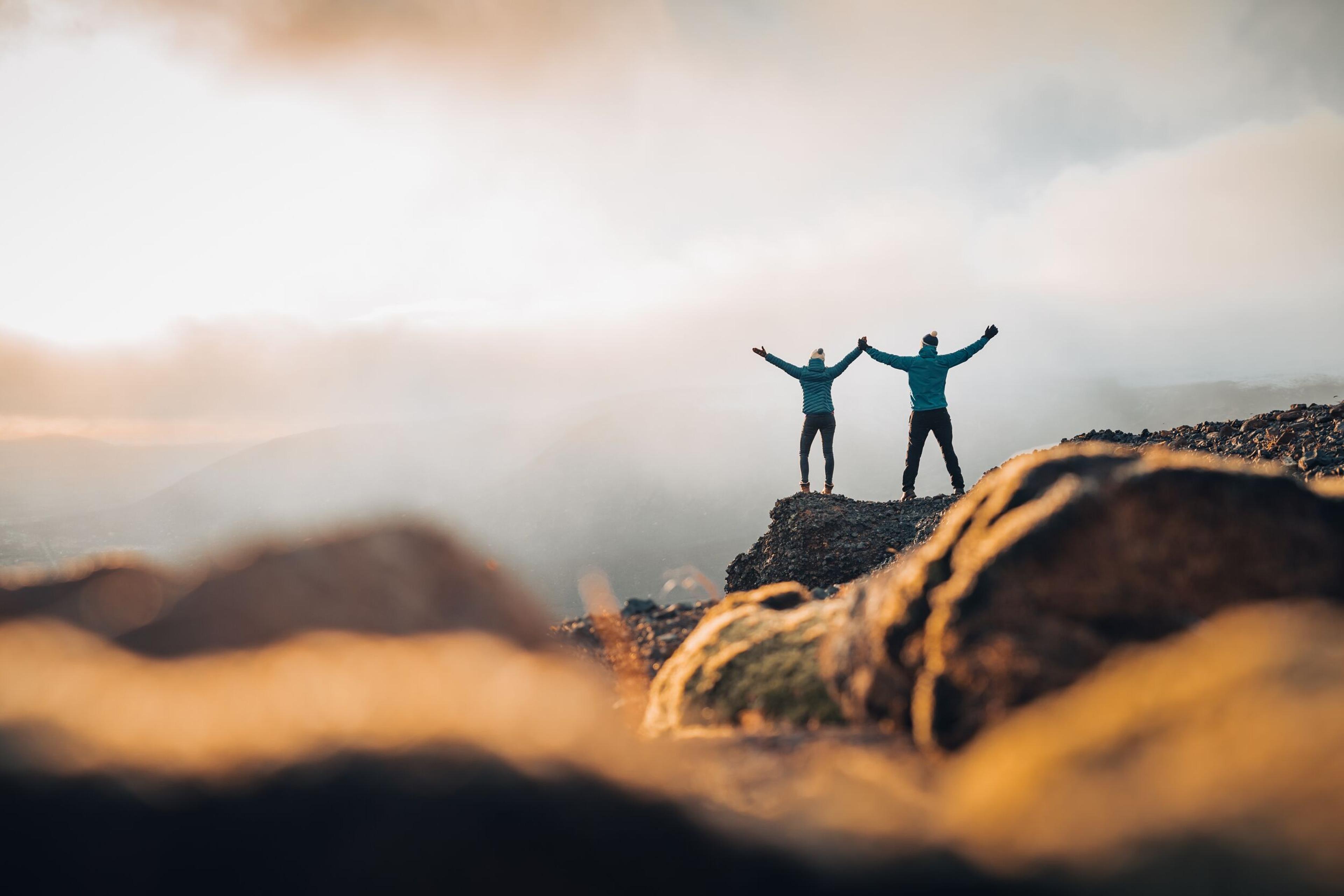 Two people stand on a mountaintop with arms raised in triumph, overlooking a cloudy landscape.