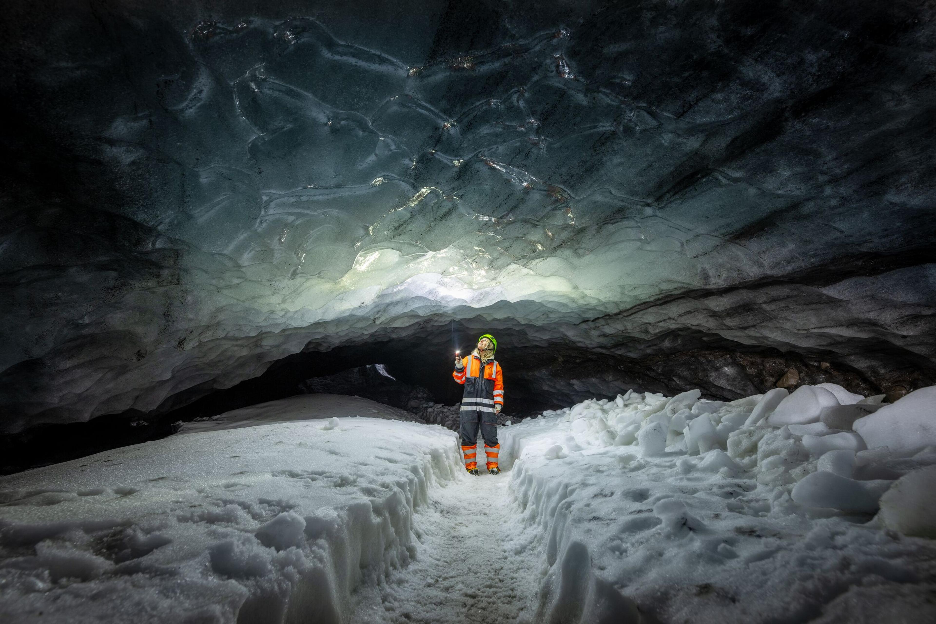 Person exploring an ice cave in Iceland, wearing an orange and black suit, surrounded by ice and snow with a glowing ceiling.