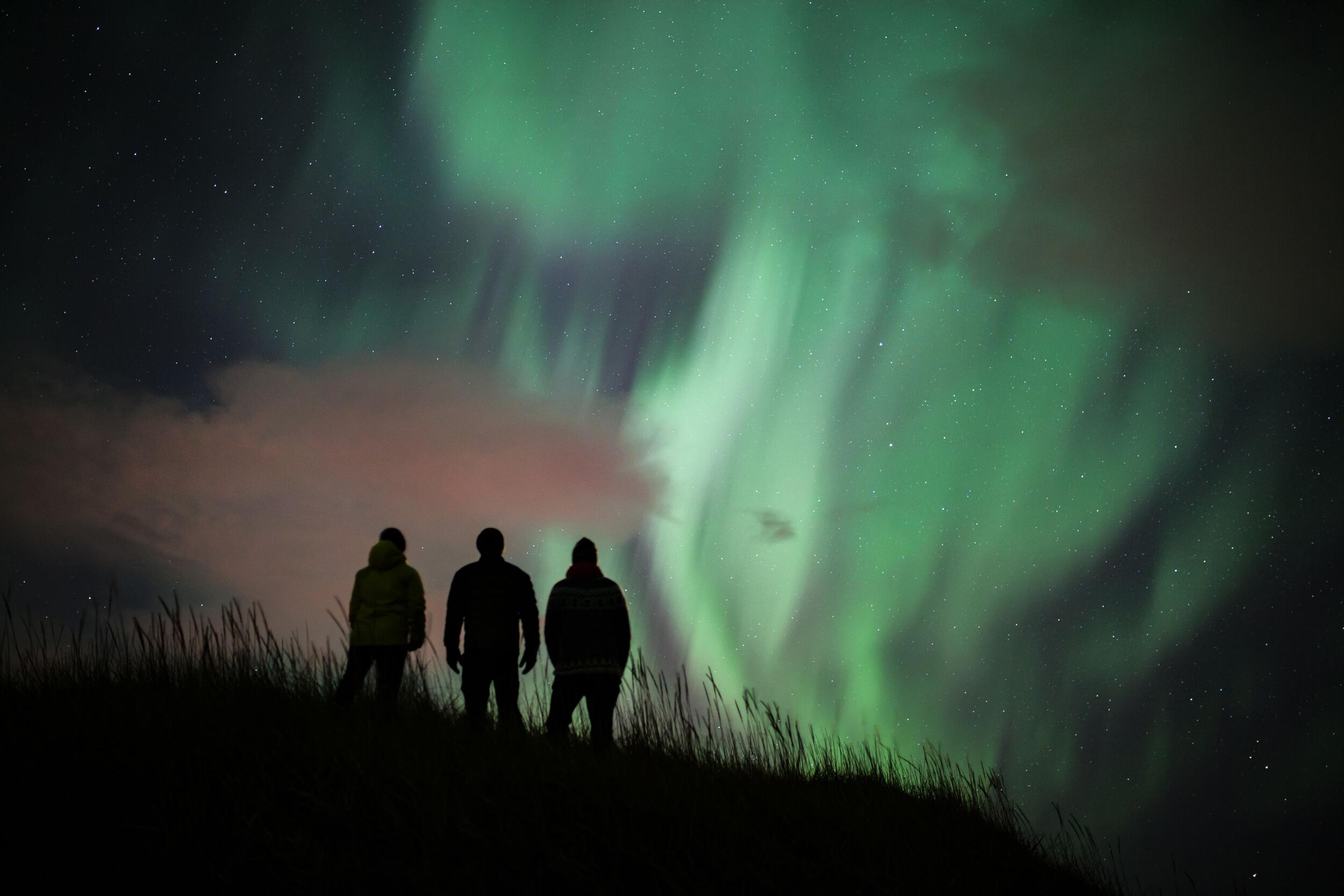 Three silhouetted figures stand against a night sky, observing the magnificent Northern Lights.