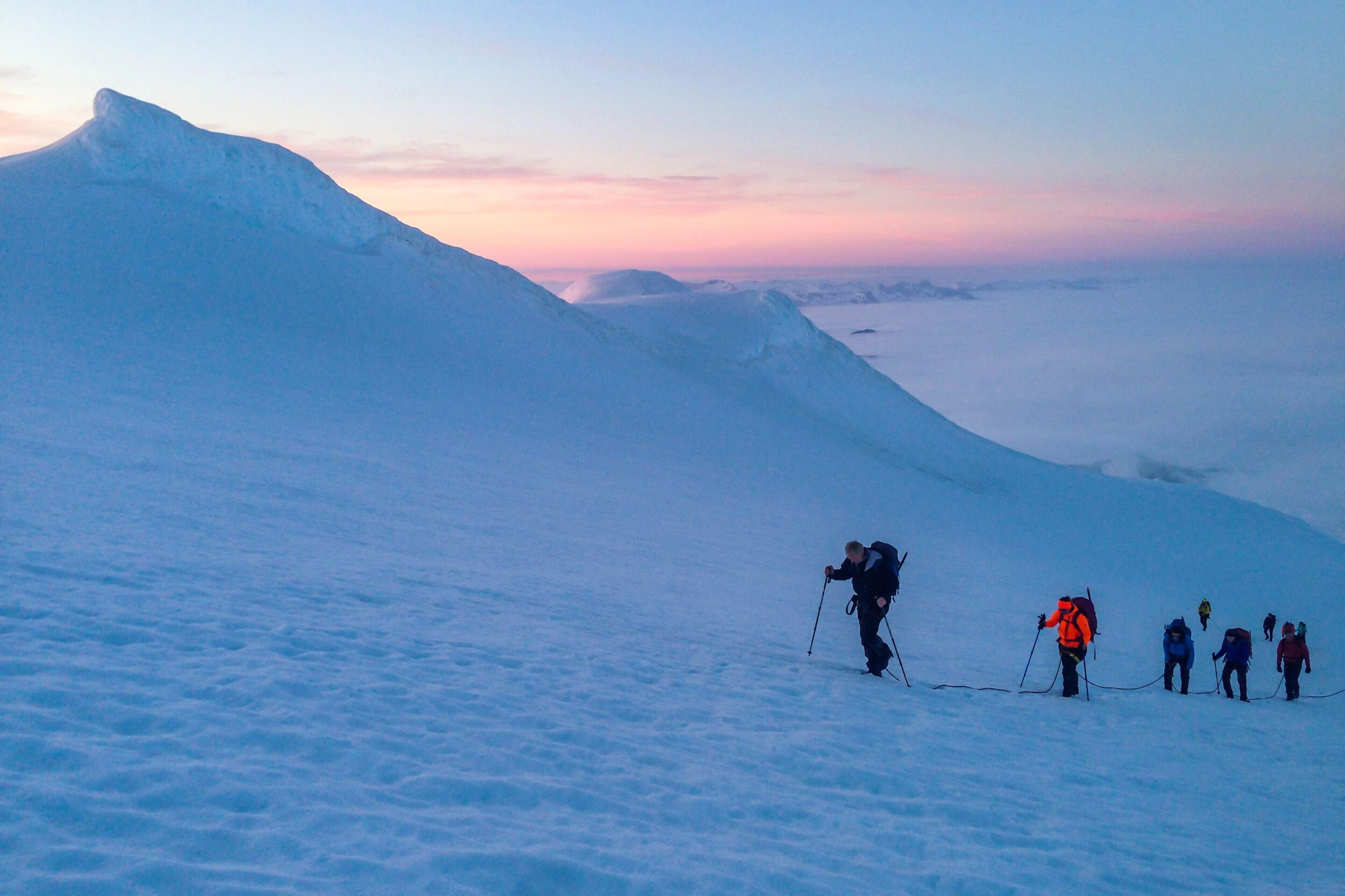 Hikers equipped with poles ascend a snowy slope against a pastel sunset sky, with the silhouette of a mountain ridge extending into the distance.